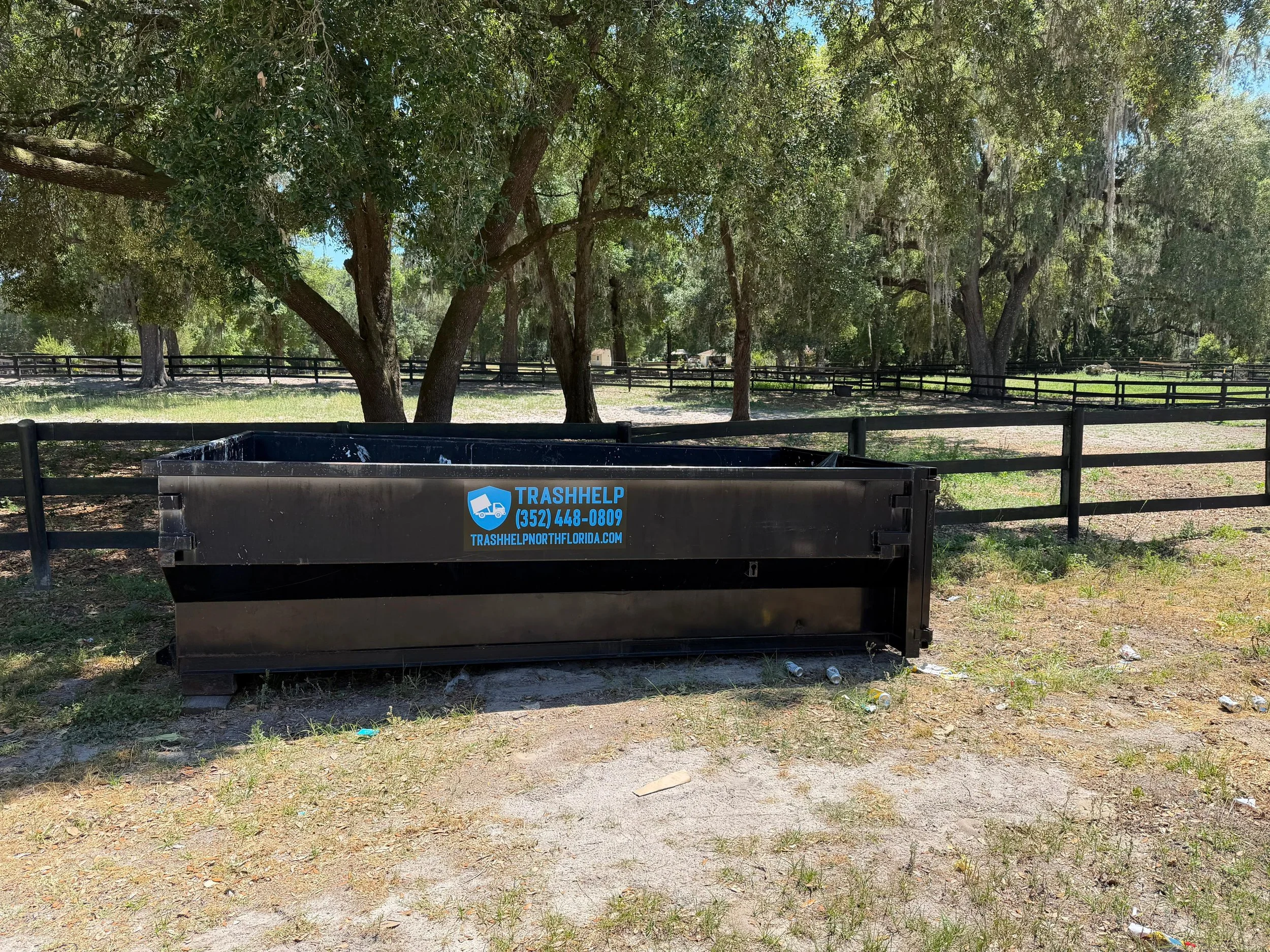 A trash container with a logo and website on it, placed outdoors on a patch of grass and dirt, with trees and a fence in the background.