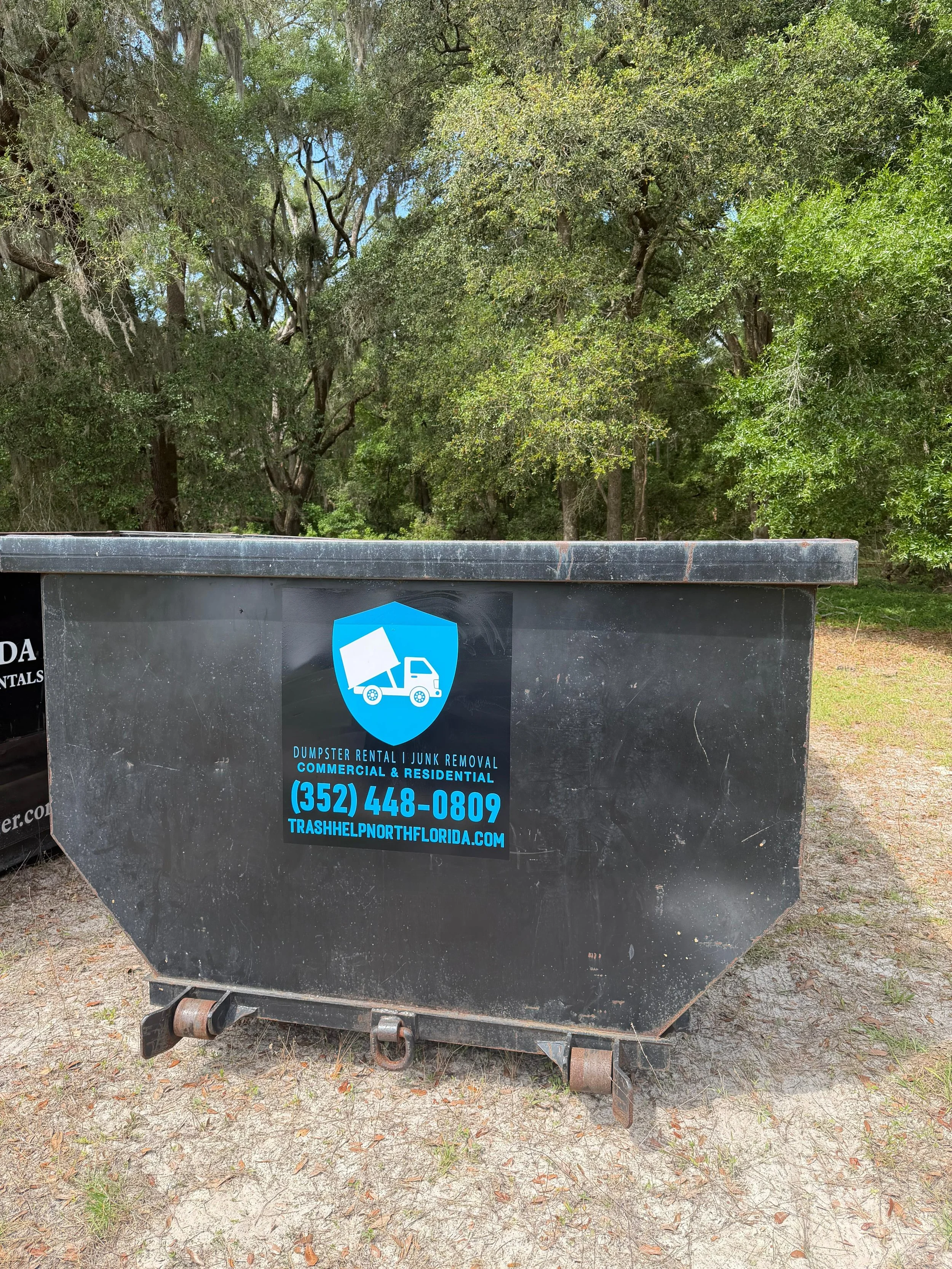 Black dumpster with a blue logo for Dumpster Rental and Junk Removal services, parked on a dirt ground with trees in the background.