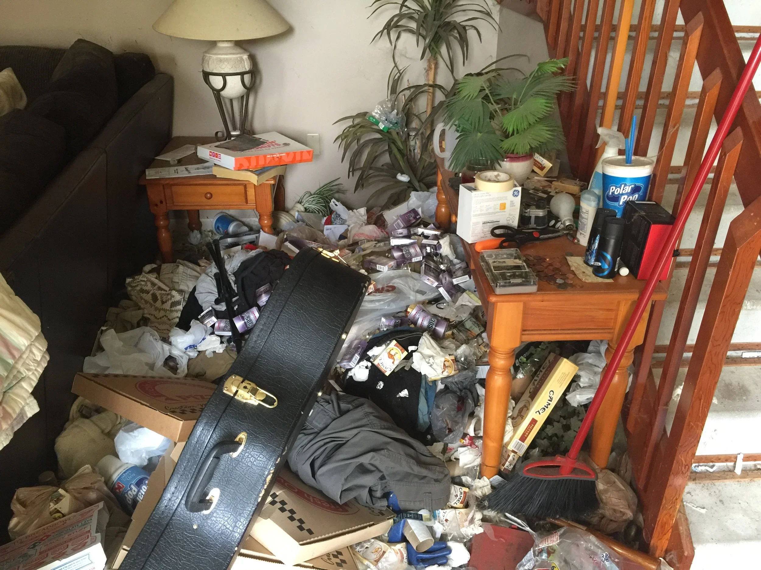 A cluttered living room corner with a black sofa, a wooden end table stacked with magazines, and a small table with various objects. The floor and nearby surfaces are covered with trash, empty bottles, paper, and miscellaneous items. There are large indoor plants and a staircase railing in the background.