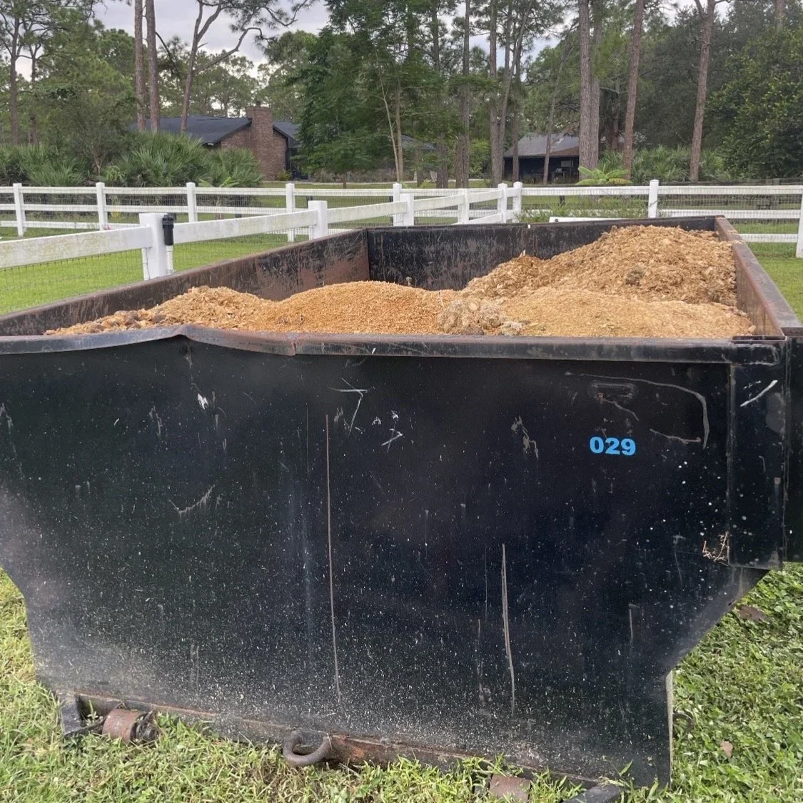 Large black dumpster filled with dirt and soil, with a rural background of trees, houses, and a white fence.