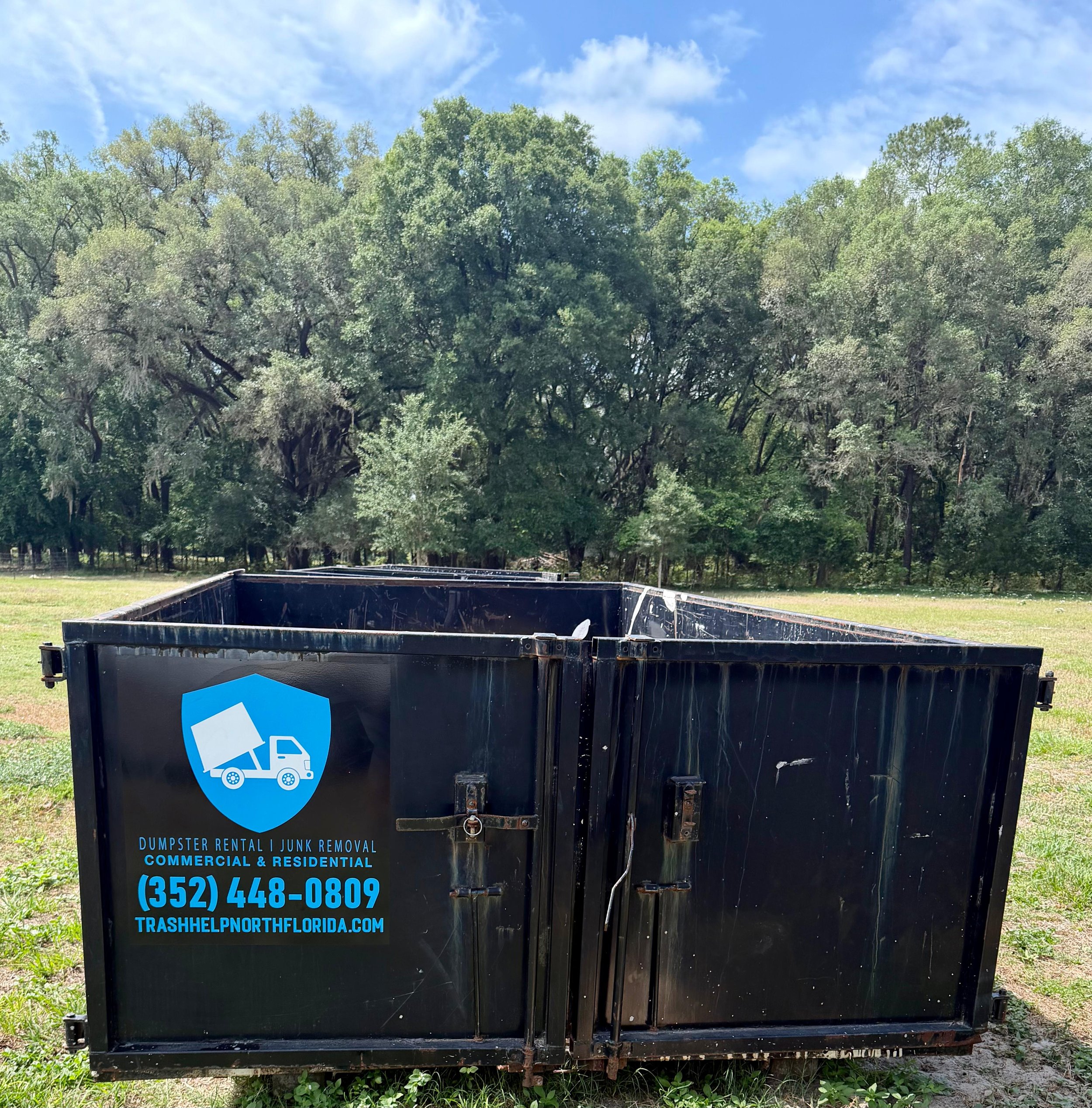 Black dumpster with a blue and white recycling logo, placed on grass with trees and a blue sky with clouds in the background.