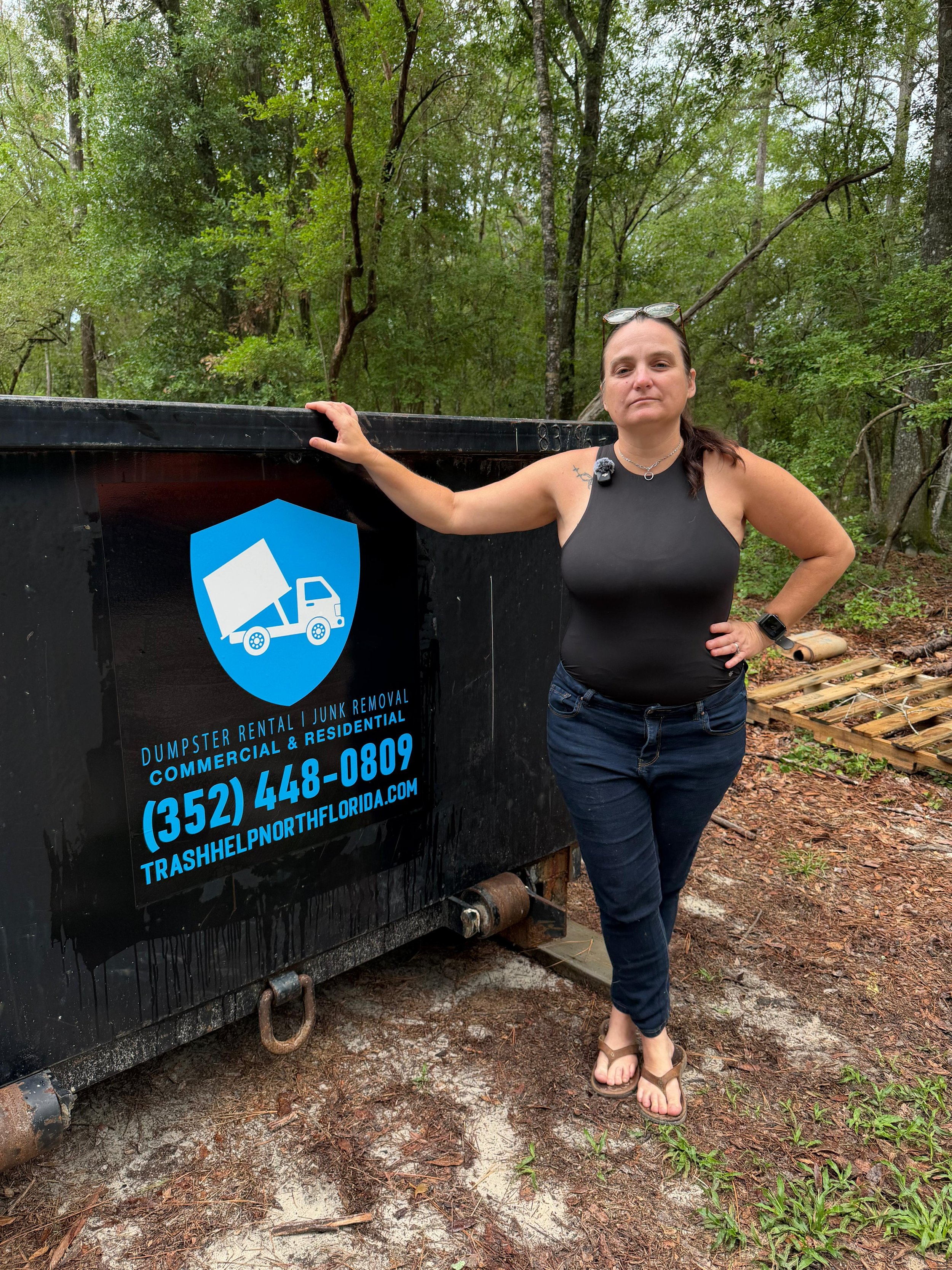 A woman wearing a black tank top, jeans, and sandals stands outdoors in front of a large black dumpster with a blue and white trash removal logo. She has her left hand on her hip and her right hand resting on the dumpster, with sunglasses on her head