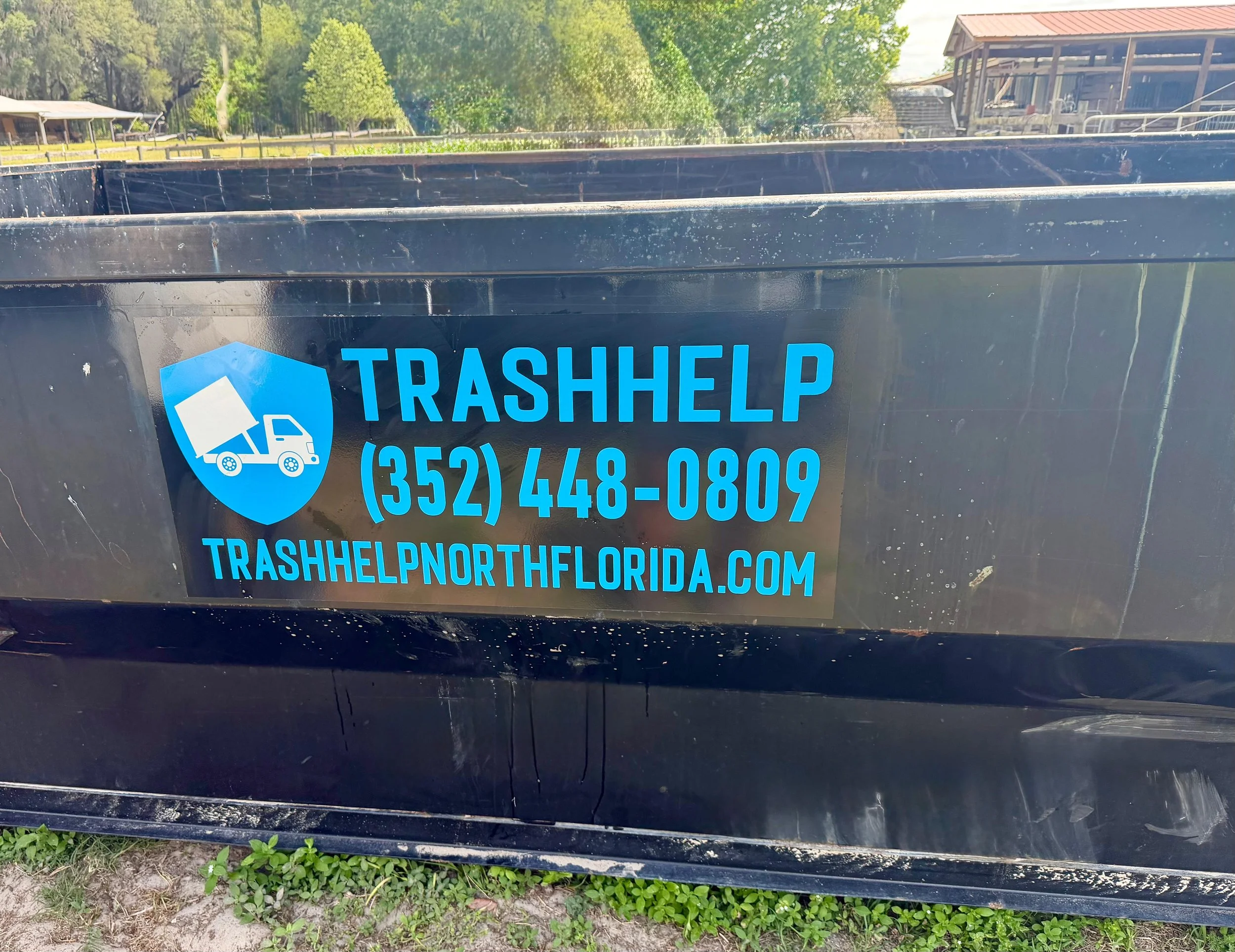 A black trash dumpster with blue lettering that reads 'TRASHHELP' and a phone number, located outdoors with trees, grass, and a building in the background.