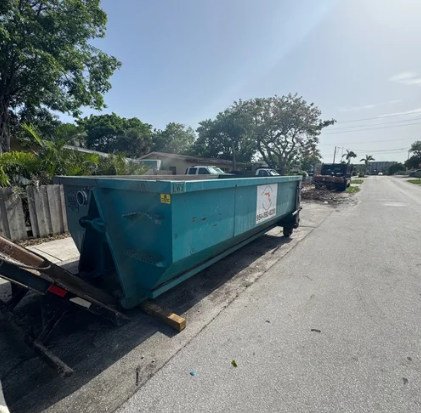 Large turquoise dumpster on a paved street with trees and houses in the background.