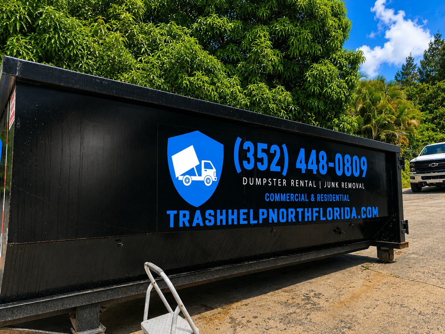 Black dumpster trailer with blue and white logo and text for TrashHelp North Florida, including phone number (352) 448-0809, services for dumpster rental and junk removal, and website trashhelpnorthflorida.com. The trailer is outdoors with green trees and a blue sky in the background.