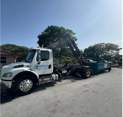 A white trash truck with a green dumpster attached, parked on a paved street in a suburban area with trees and houses.