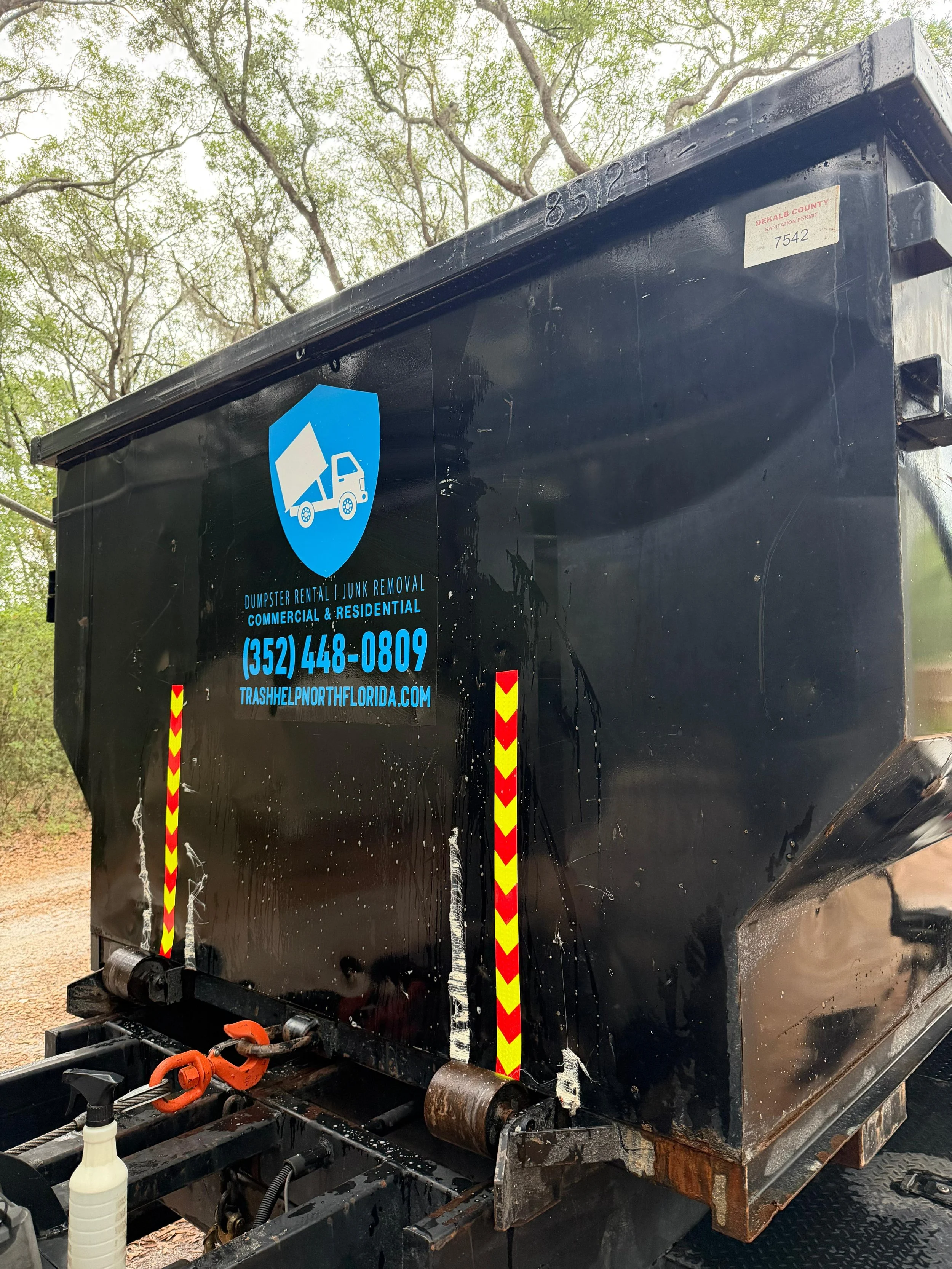 Black dumpster with a blue logo depicting a truck and trash container, and contact info including phone number and website, outdoors with trees in the background.