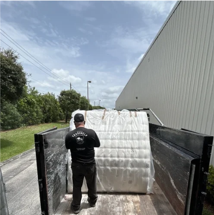 A man in black clothing standing next to a large wrapped mattress inside the bed of a truck parked outdoors near a large building.