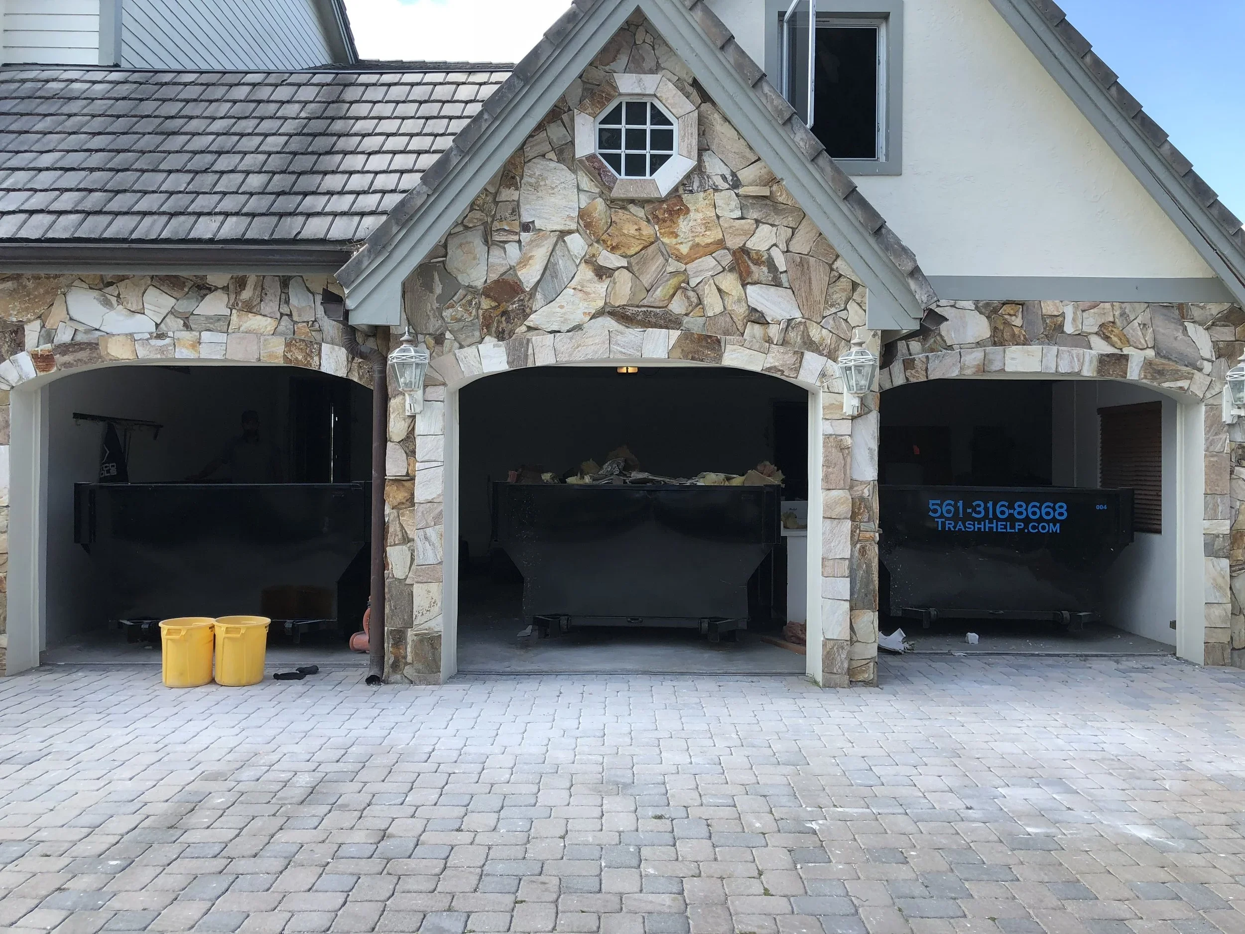 Photo of a house with a stone and stucco facade and three garage doors. Two yellow trash bins are in front of the left garage, and debris is inside the open garages. The house has a tile roof and a small octagonal window above the middle garage.