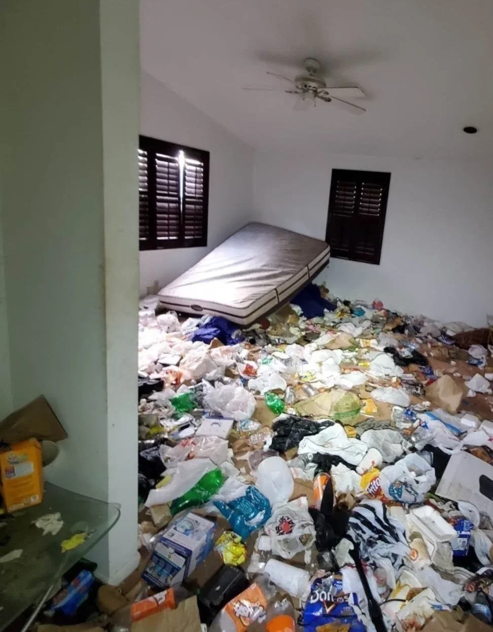 Room filled with trash, an unmade bed tilted on the floor, windows with closed blinds, ceiling fan, cluttered and messy environment.