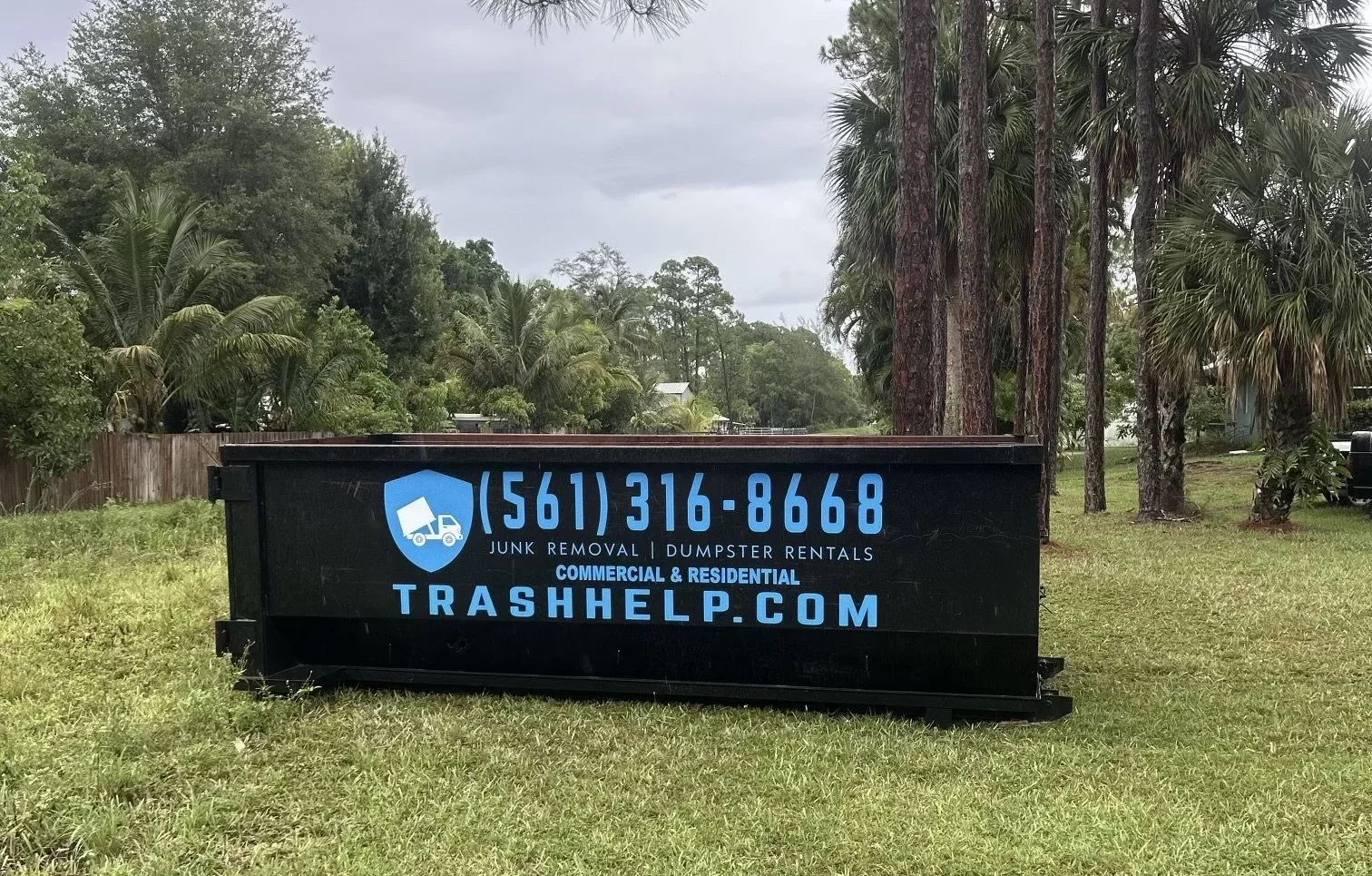 A black dumpster with blue and white lettering advertising trash removal services, including phone number, website, and services for commercial and residential junk removal and dumpster rentals, set in a grassy yard with trees and a wooden fence in t