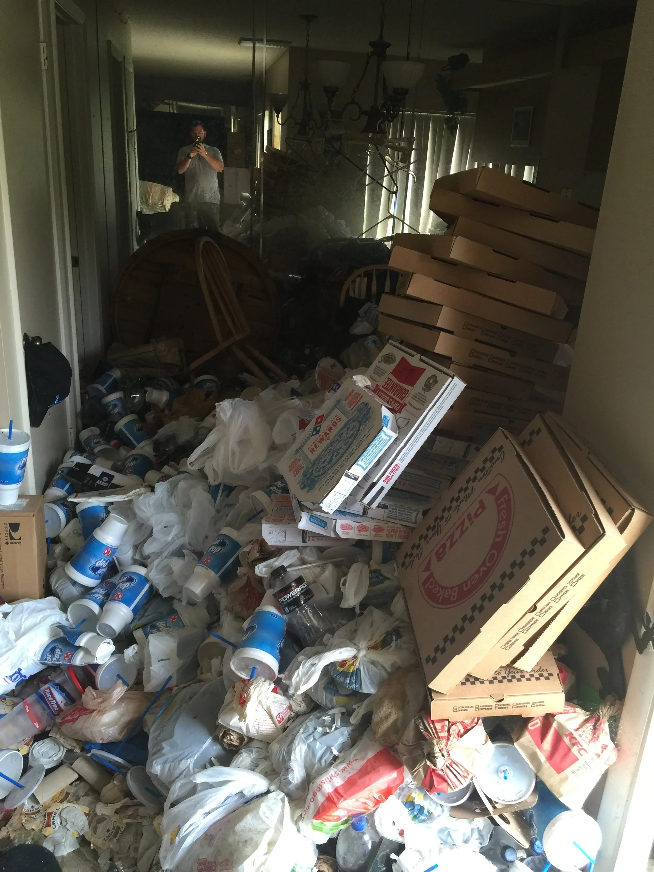 A cluttered room filled with discarded paper bags, pizza boxes, plastic bottles, and miscellaneous trash, with a person taking a photo of the mess in a mirror.