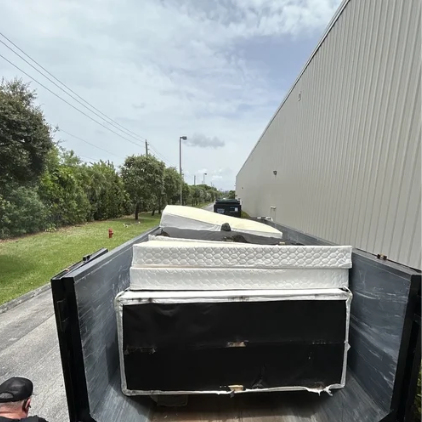 View from the back of a flatbed truck carrying large rectangular metallic objects, parked alongside a building with trees and power lines in the background.