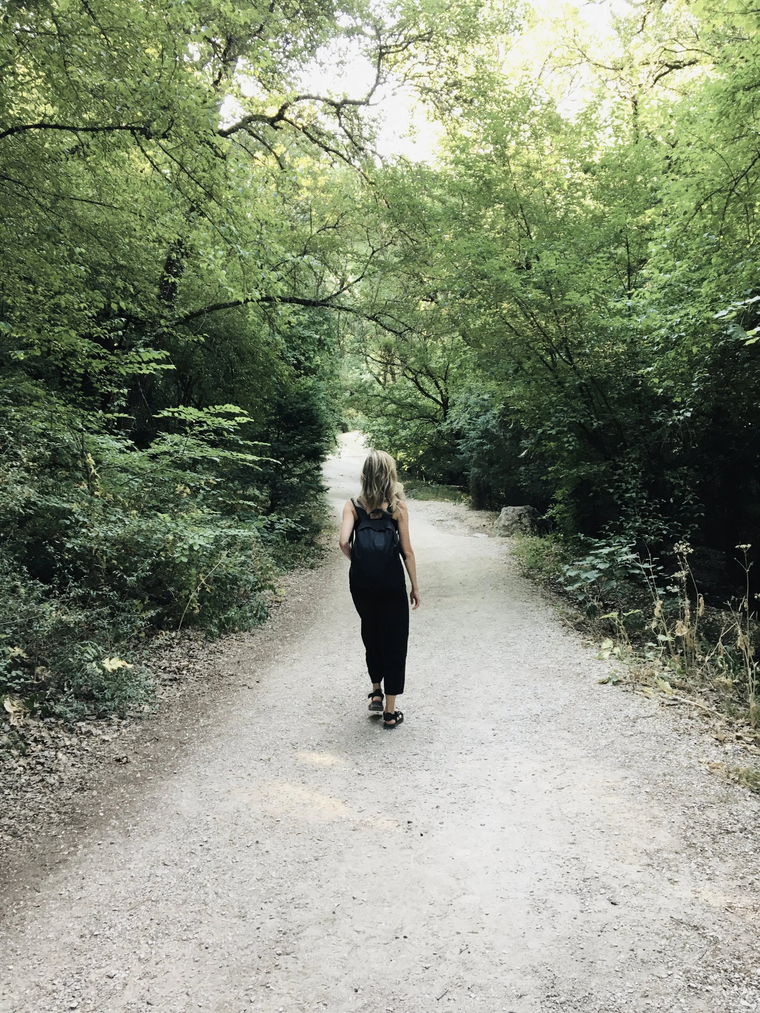 A woman with blonde hair, wearing a black outfit and a backpack, walking down a dirt path surrounded by green trees and foliage.
