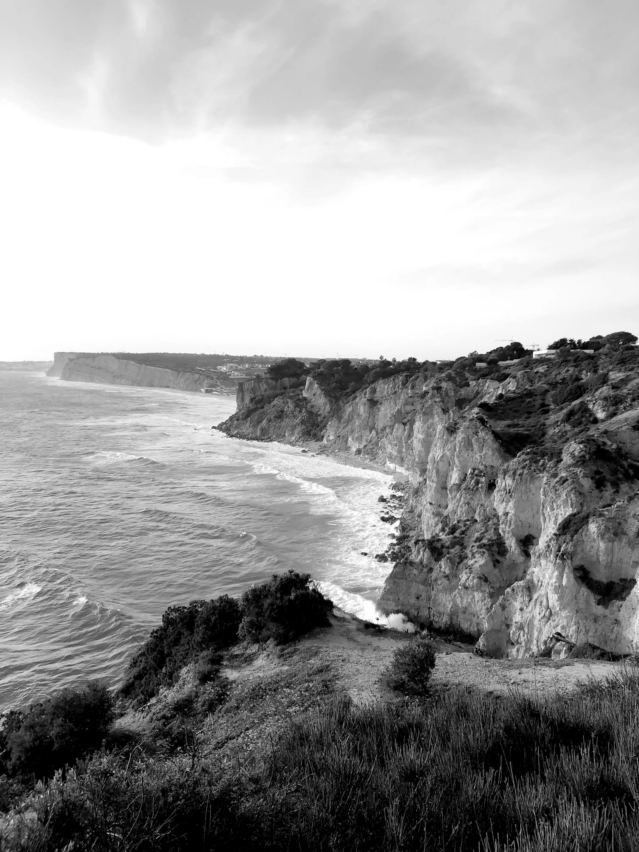 Black and white photograph of rugged cliffs along the coast with ocean waves crashing at the base, distant land on the horizon, and a cloudy sky overhead.
