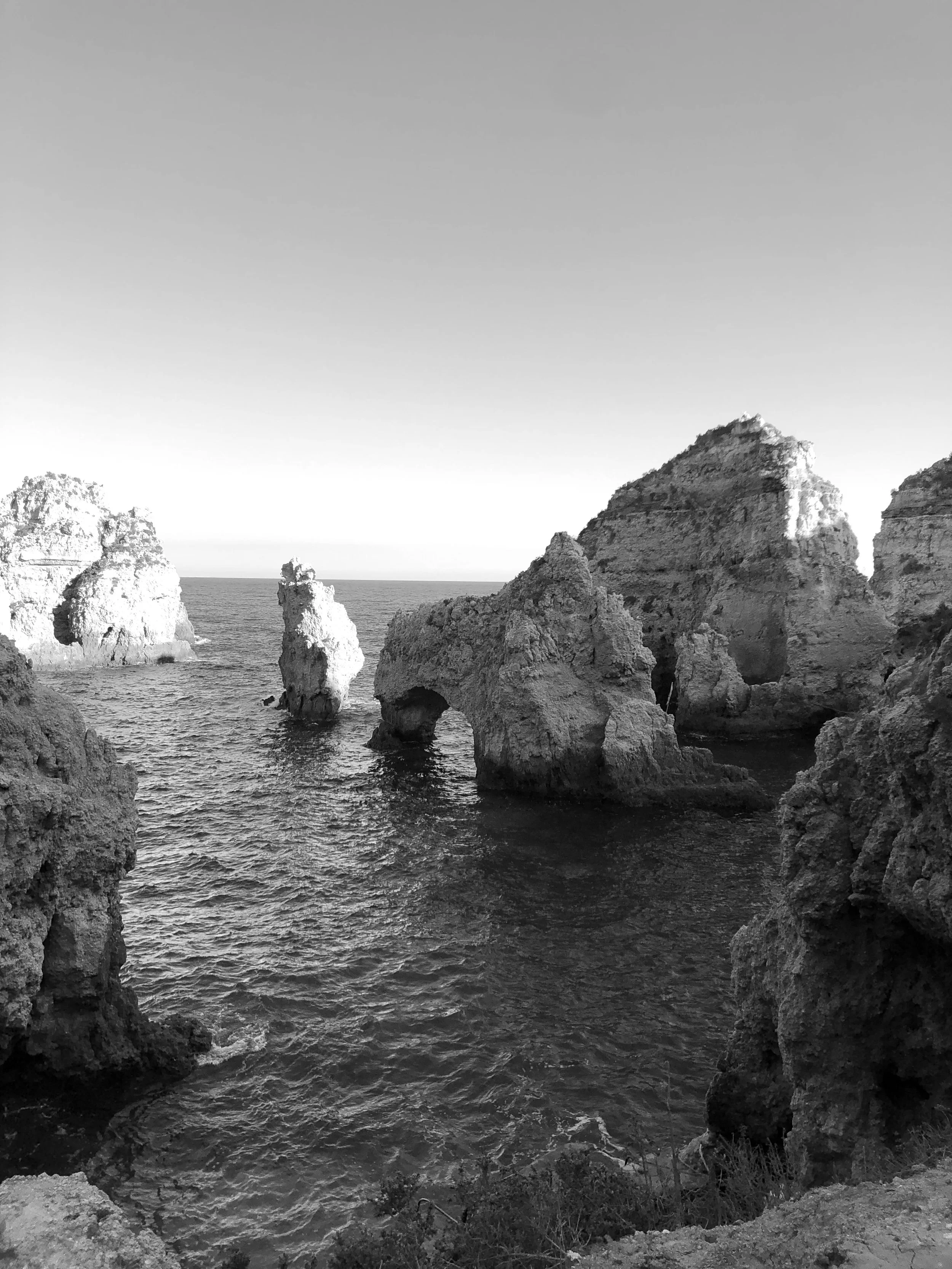 Black and white photograph of rocky coastal cliffs and sea stacks at the ocean with calm water and clear sky.