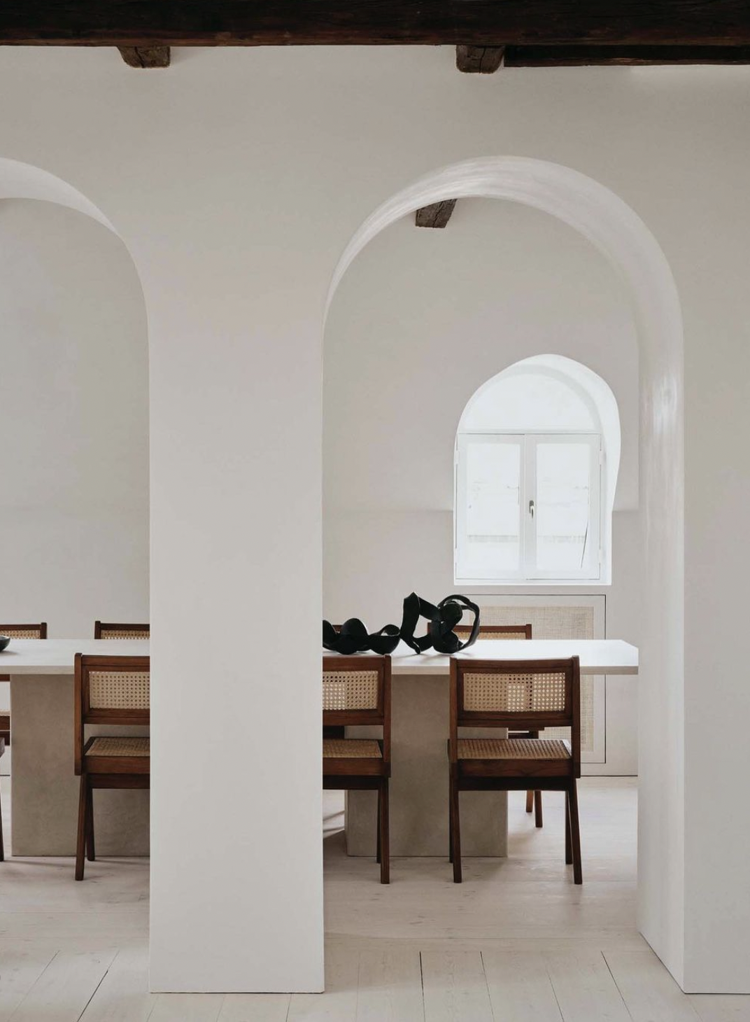 Minimalist dining area with wooden chairs around a white table, black headphones resting on the table, arched windows, white walls, and wooden ceiling beams.