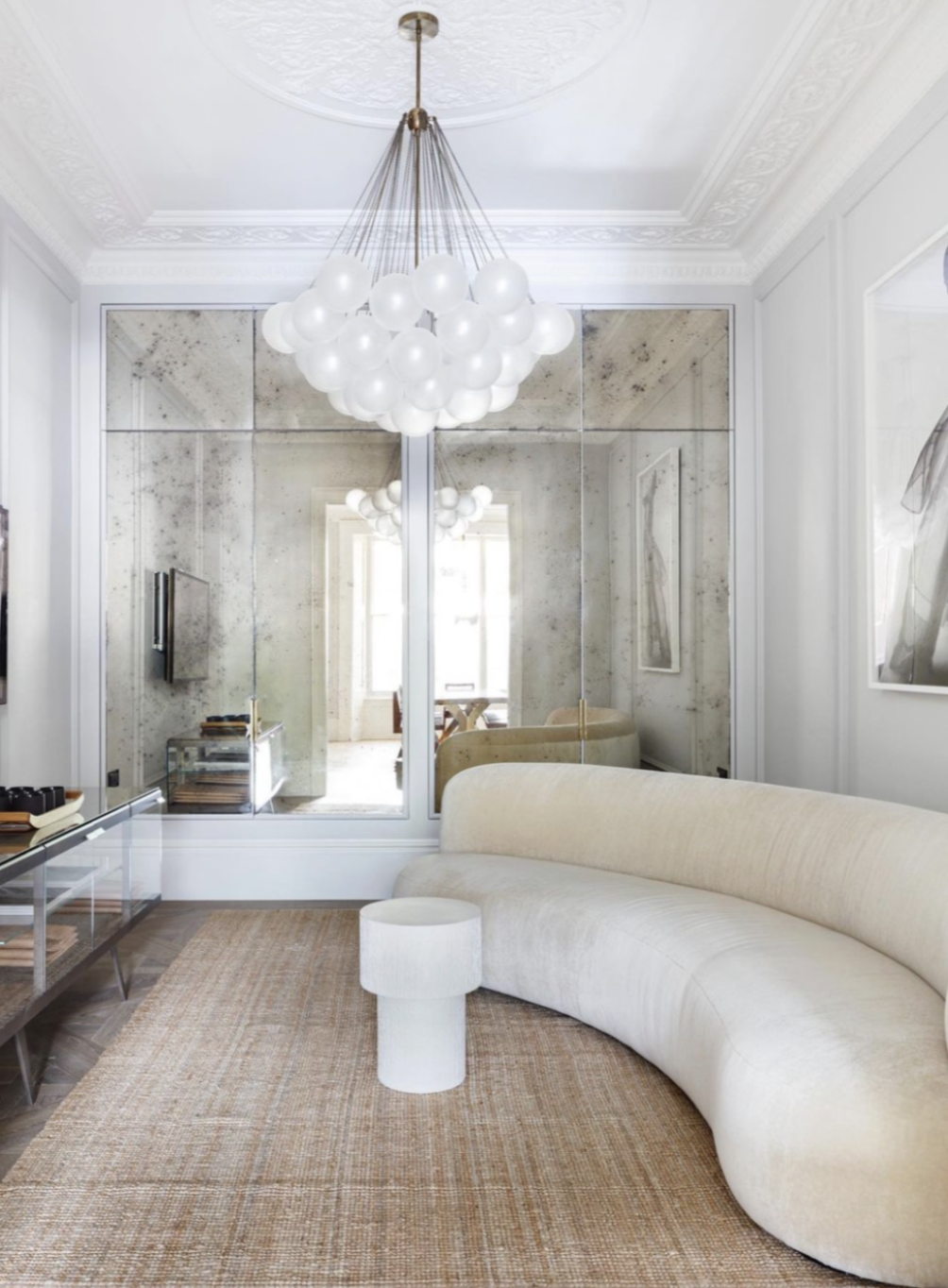 Living room with ornate ceiling, white walls, mirrored panel, modern chandelier with white globes, curved beige sofa, small white round side table, and large framed art.
