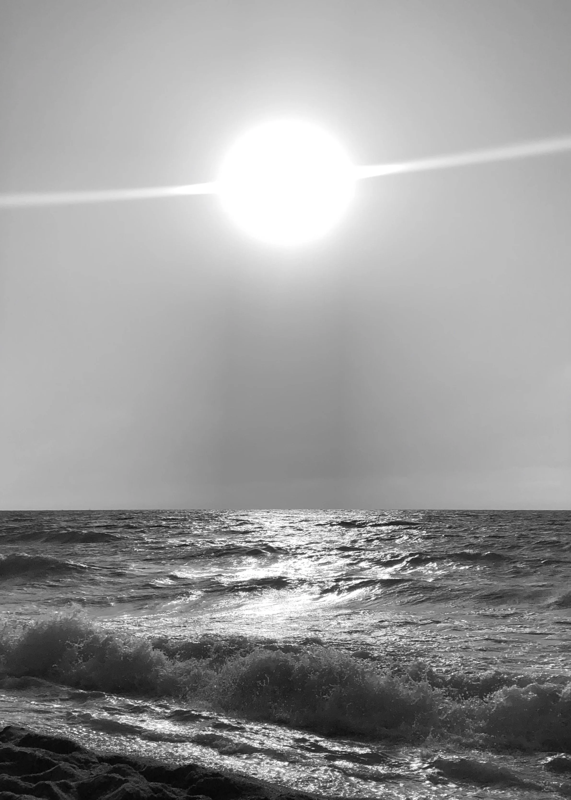 Black and white photo of sunlight reflecting off the ocean waves, with the sun positioned high in the sky.