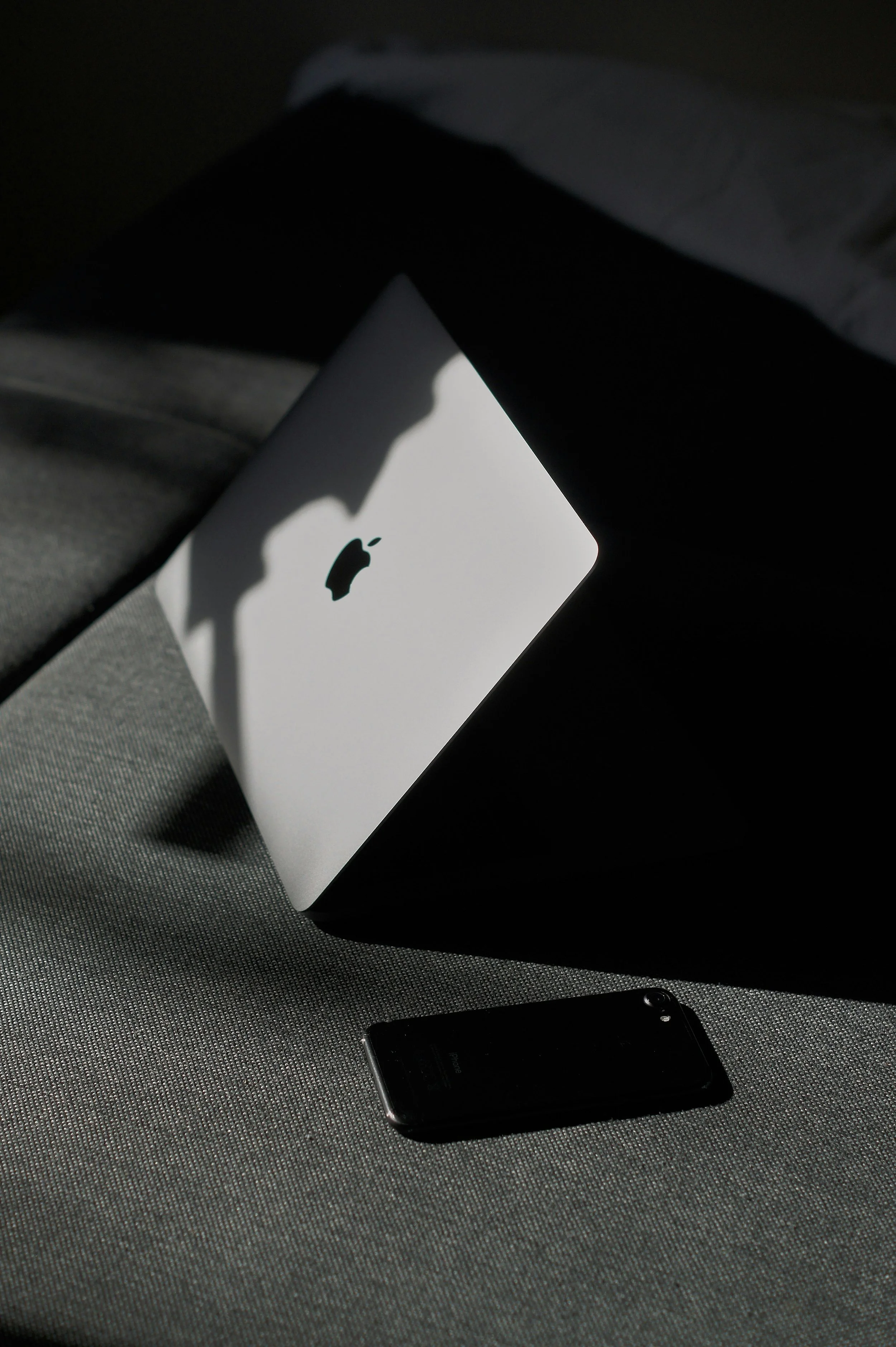 An Apple MacBook laptop and an iPhone placed on a textured surface with shadows cast across them.