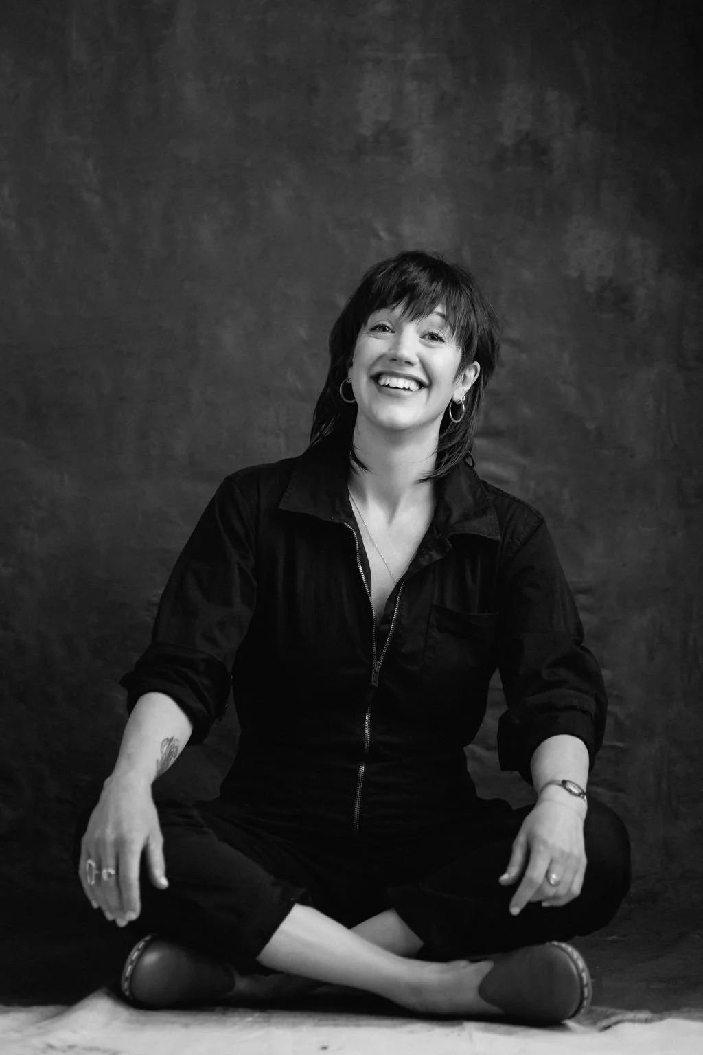 Black and white photo of a smiling woman sitting cross-legged on the floor against a dark backdrop, wearing a zip-up jacket and dark pants.