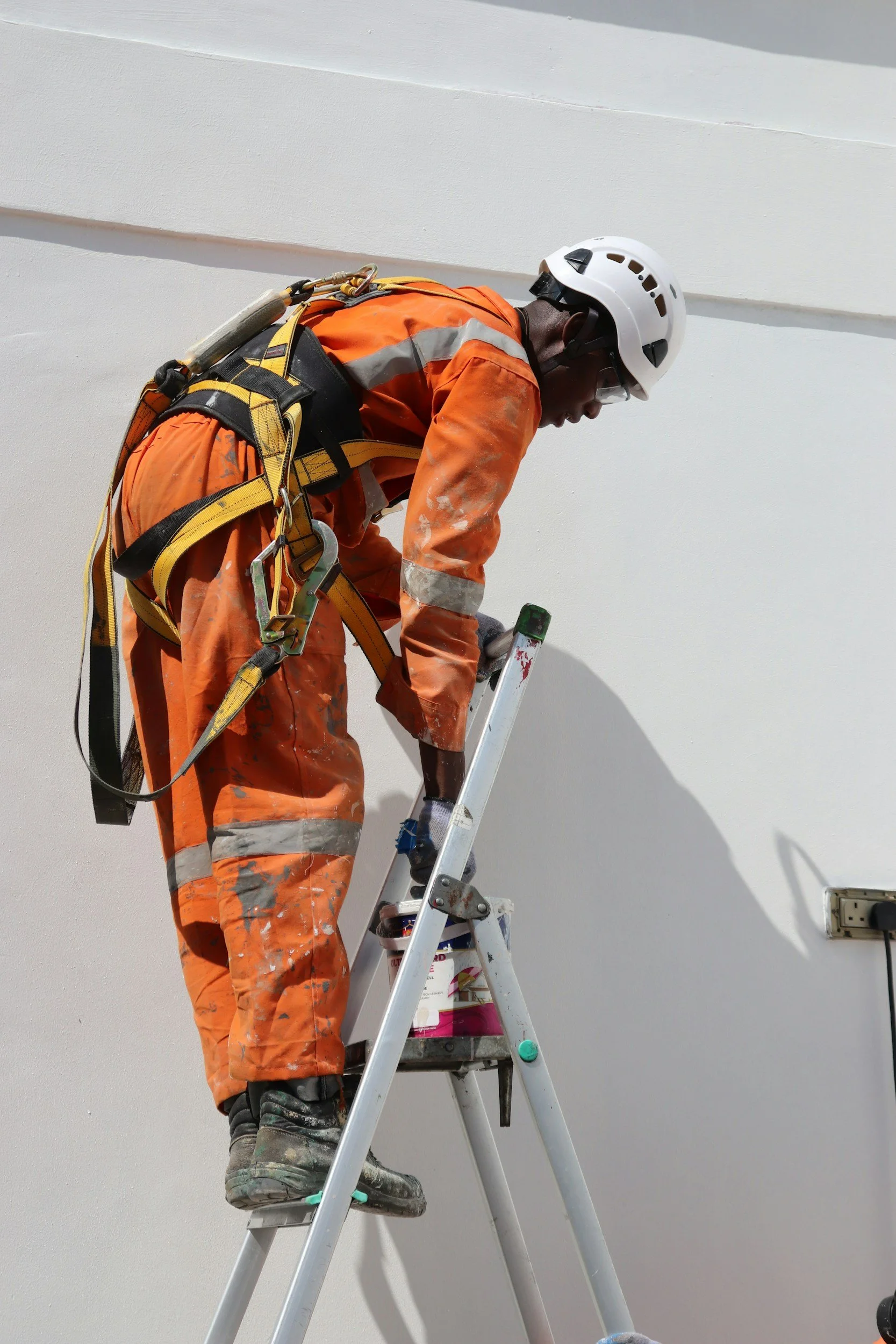 Worker in orange safety clothing and helmet standing on ladder painting a white wall.