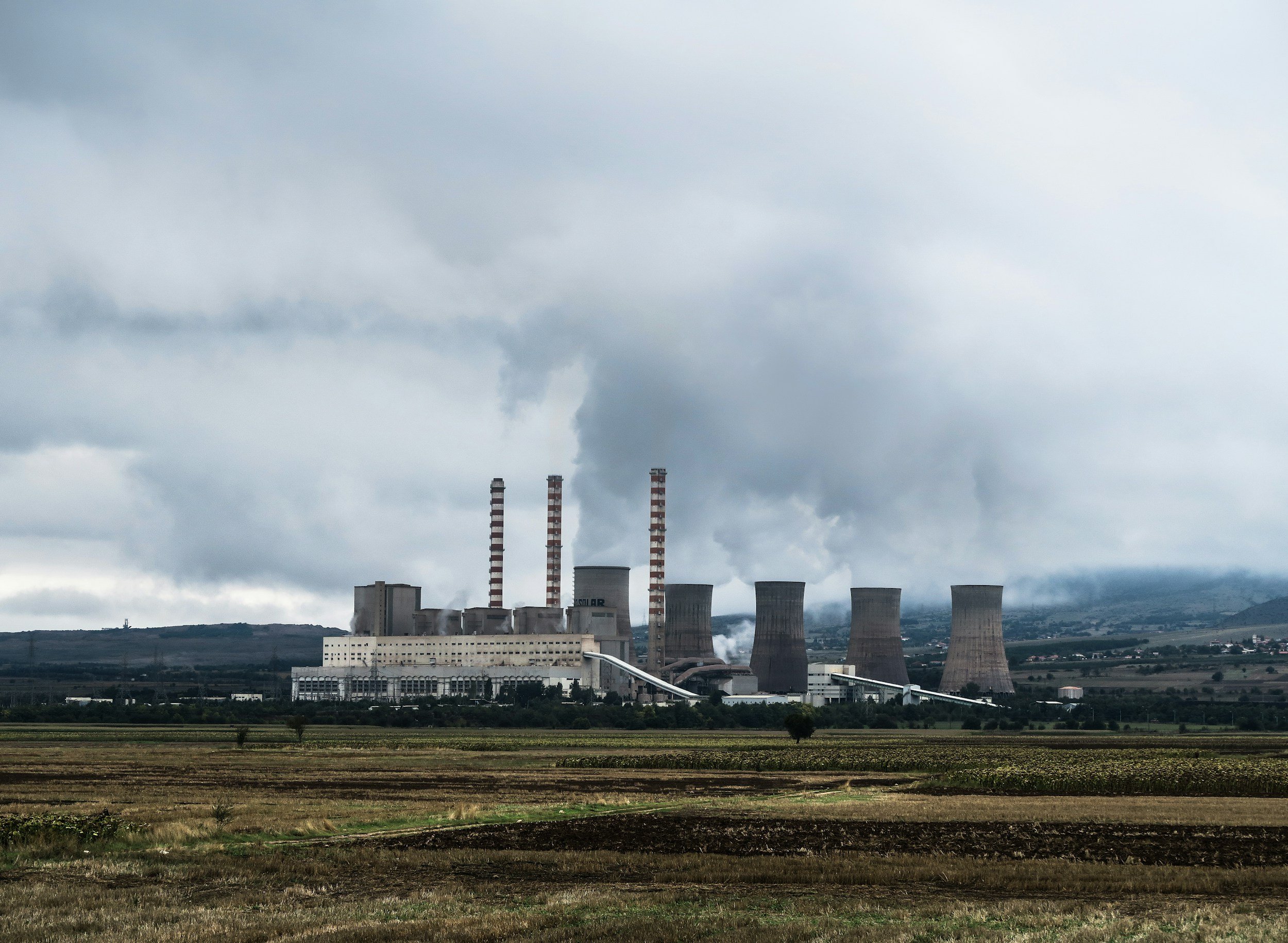 A large industrial power plant with four cooling towers and three smokestacks emitting smoke or steam, set against a cloudy sky and open farmland in the foreground.
