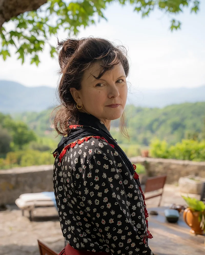 A woman with dark hair in a bun, wearing a black shirt with white and red flowers, standing outdoors with a scenic landscape of green hills and mountains in the background.