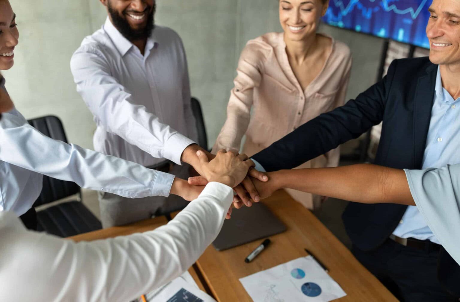 A diverse group of five professionals is gathered around a conference table in a meeting room with charts on the wall, placing their hands together in a show of teamwork and collaboration.