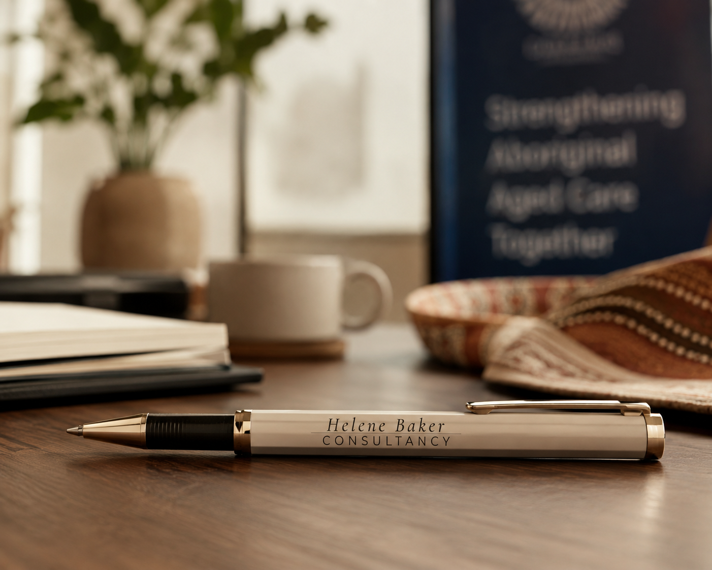 Close-up of a silver and black pen with the text 'Helene Baker CONSULTANCY' on it, resting on a wooden desk. In the background, there is a potted plant, a coffee mug, a notebook, a book, and a framed sign with partially visible text.