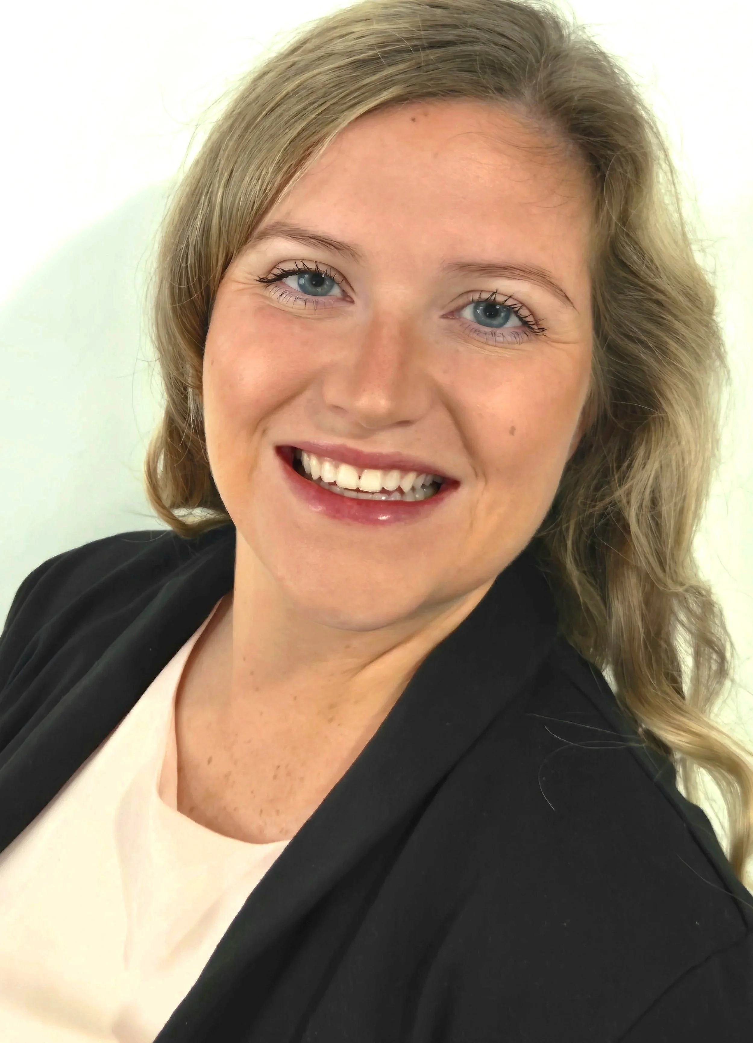 Close-up of a smiling woman with wavy blonde hair, blue eyes, wearing a black blazer and a light-colored top, against a plain white background.