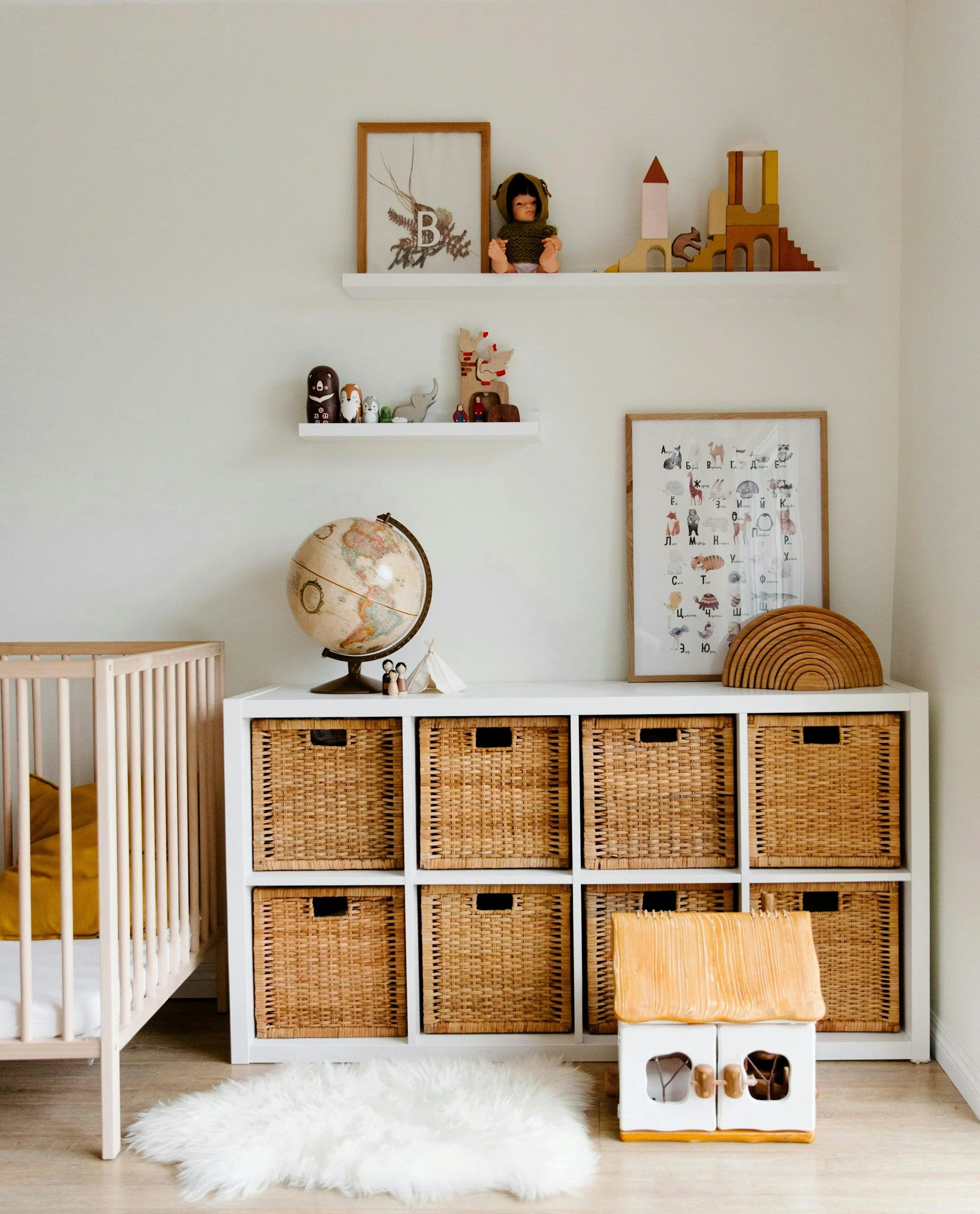 Decorative shelves with toys and artwork in a nursery room, including a globe, plush animal figures, a framed picture, a stack of woven baskets, a fluffy white rug, and a small white and wooden play structure.