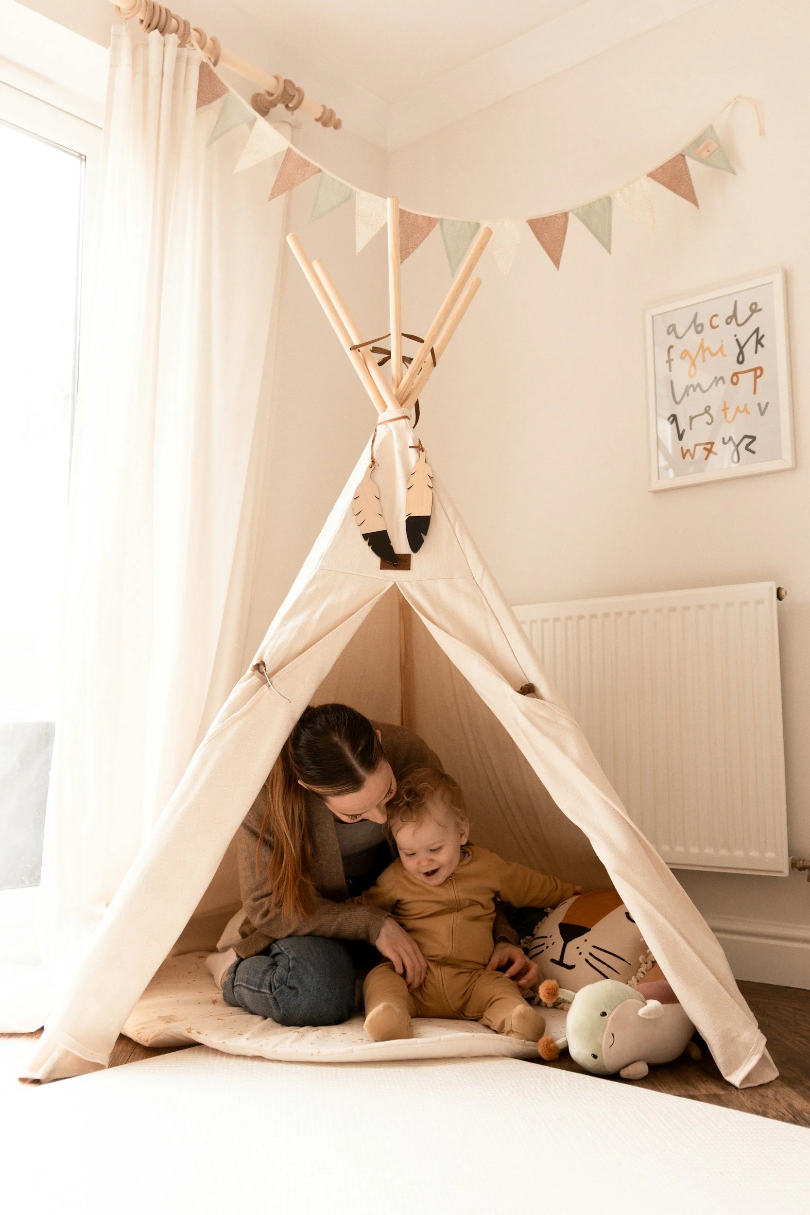 A woman and a young boy sitting inside a beige teepee tent in a well-lit room. The woman is smiling and looking at the child, who is also smiling. There is a large plush toy on the floor near them, and a soft mat on the ground.