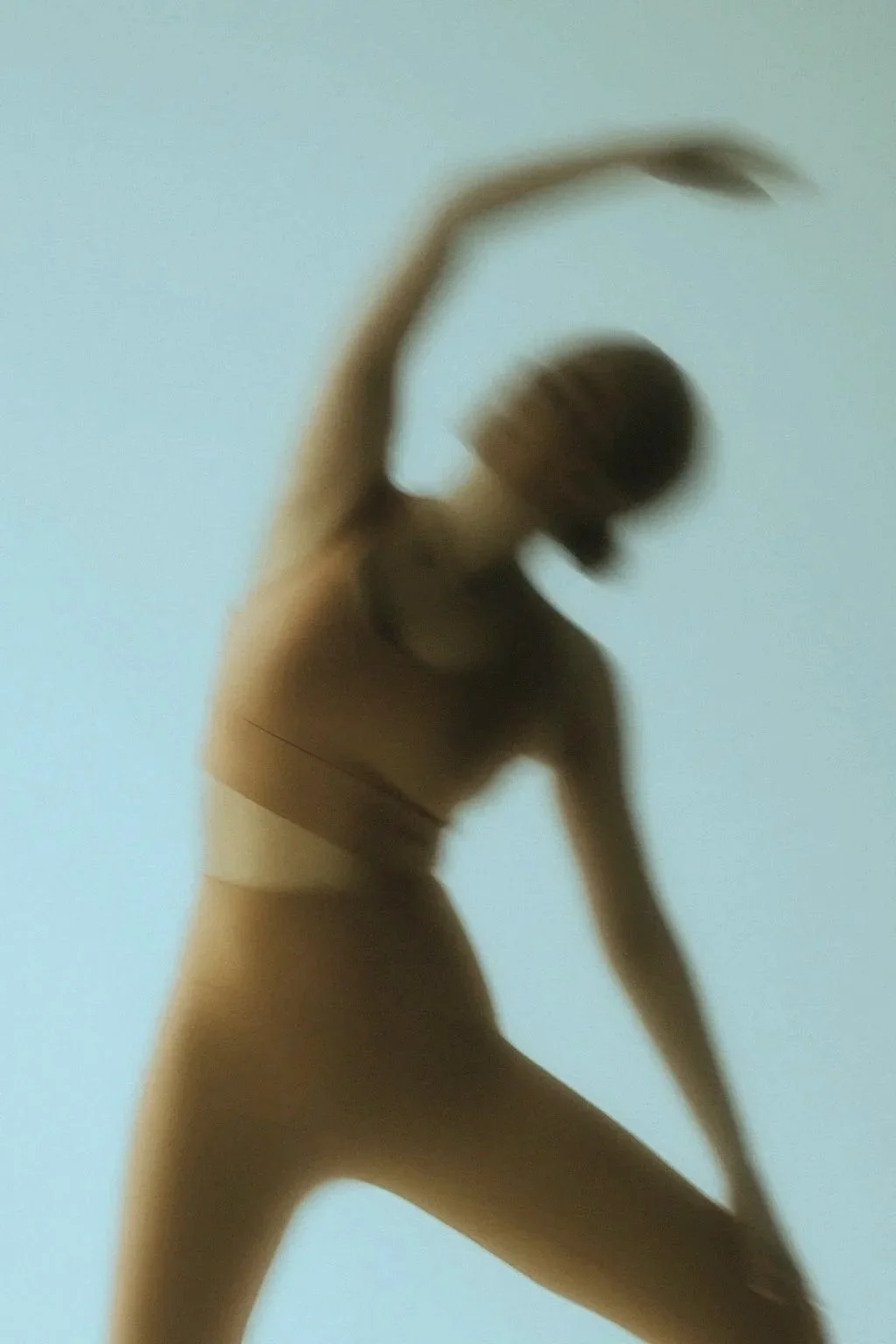 Blurred silhouette of a woman in a pose with one arm raised above her head, wearing a sports bra and leggings, against a plain background.