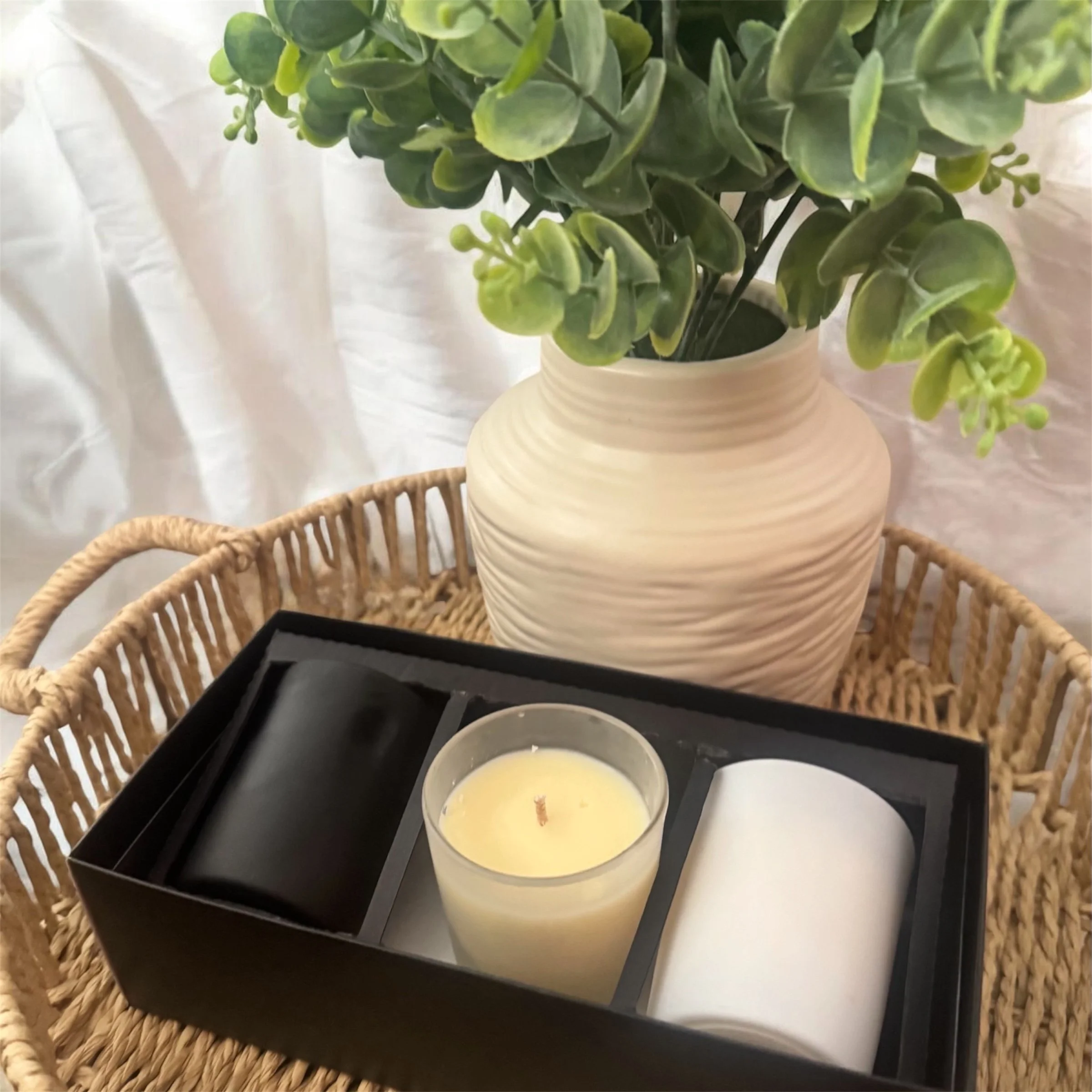 A beige vase with green and yellow artificial plants, placed on a black tray with three white candles, on a wicker table with a white curtain background.