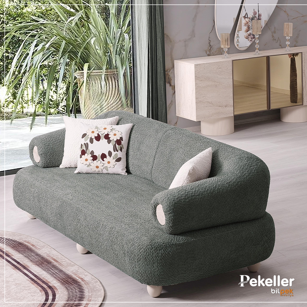 Modern living room with a green sofa, floral pillows, large glass window showing outdoor plants, and a light-colored sideboard with decorative items.