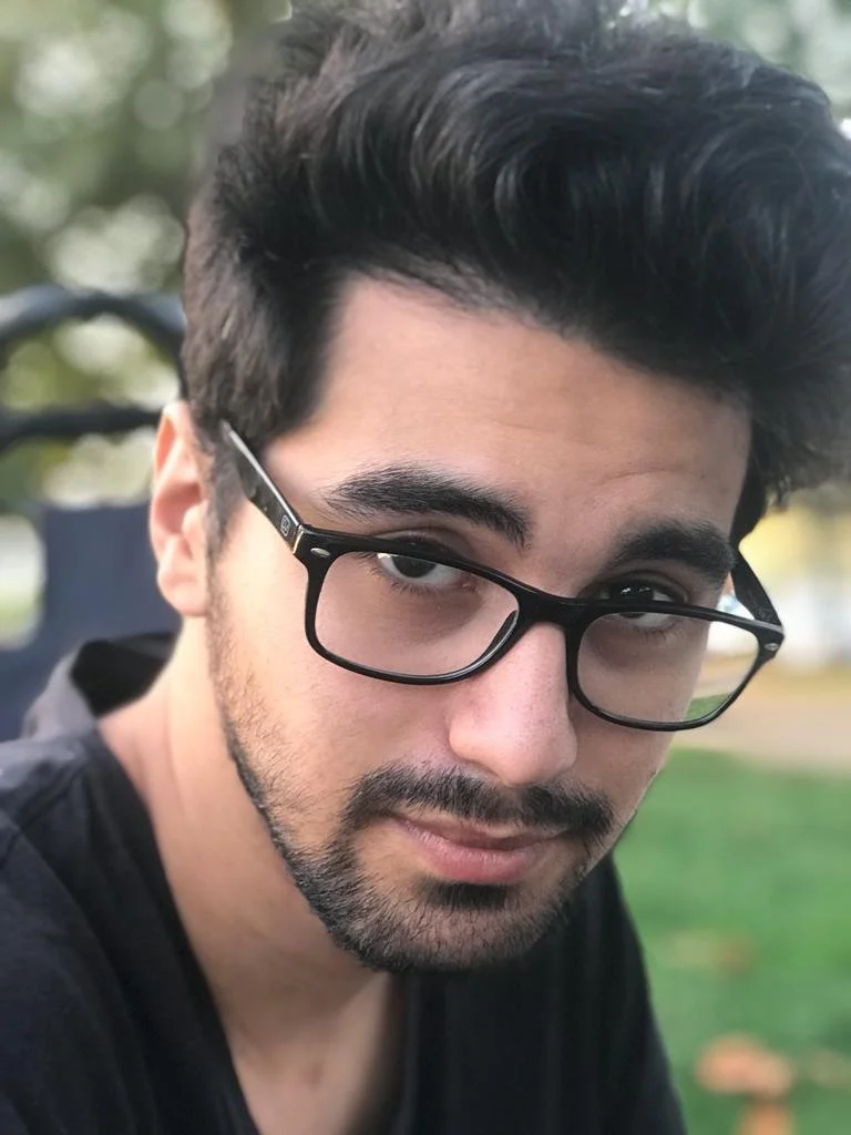 A young man with dark hair, glasses, and a beard looking at the camera outdoors with blurred trees and grass in the background.