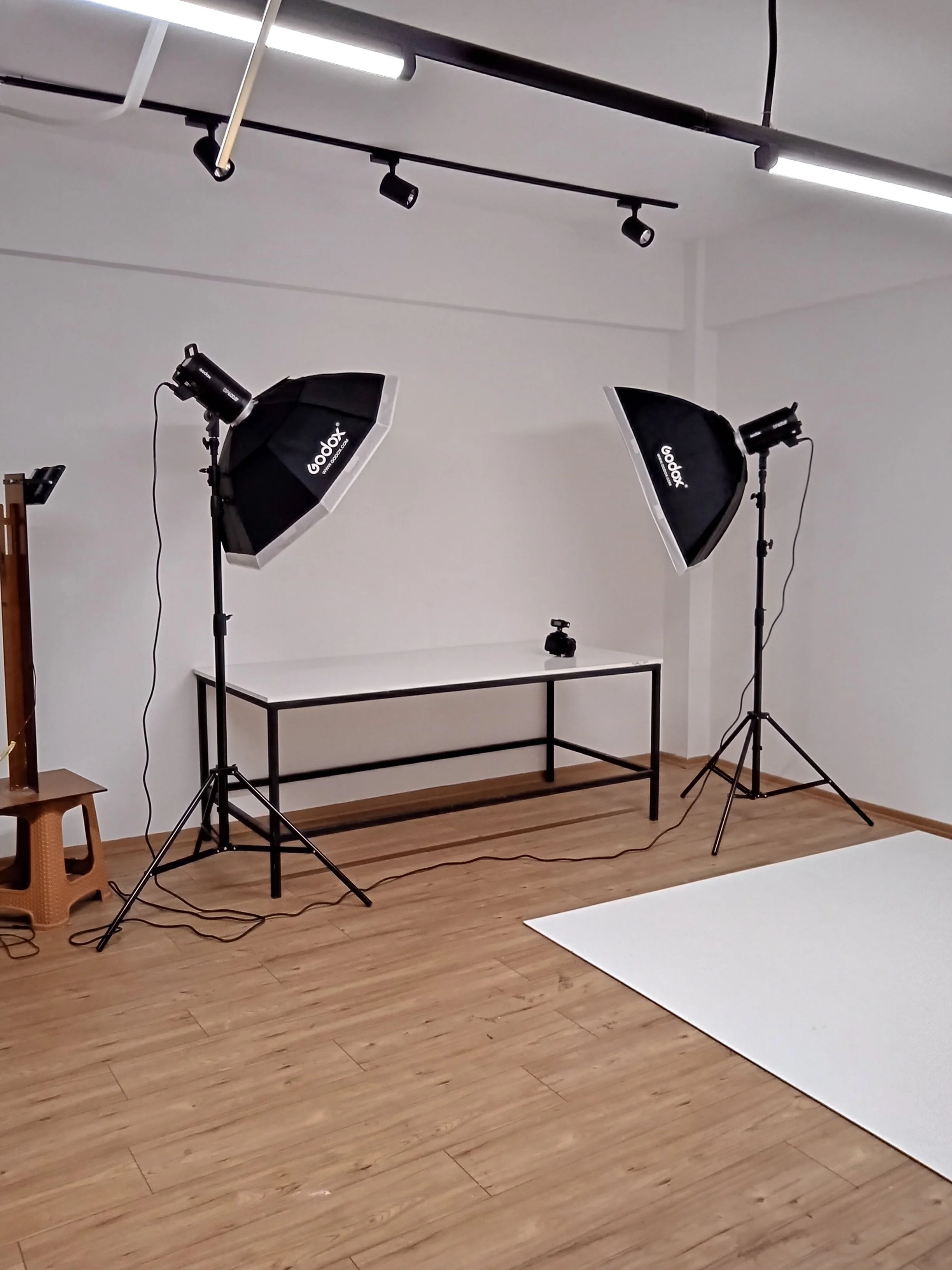 A photography studio setup with two softboxes, a camera on a table, and studio lighting, in a room with wooden flooring and a white backdrop.