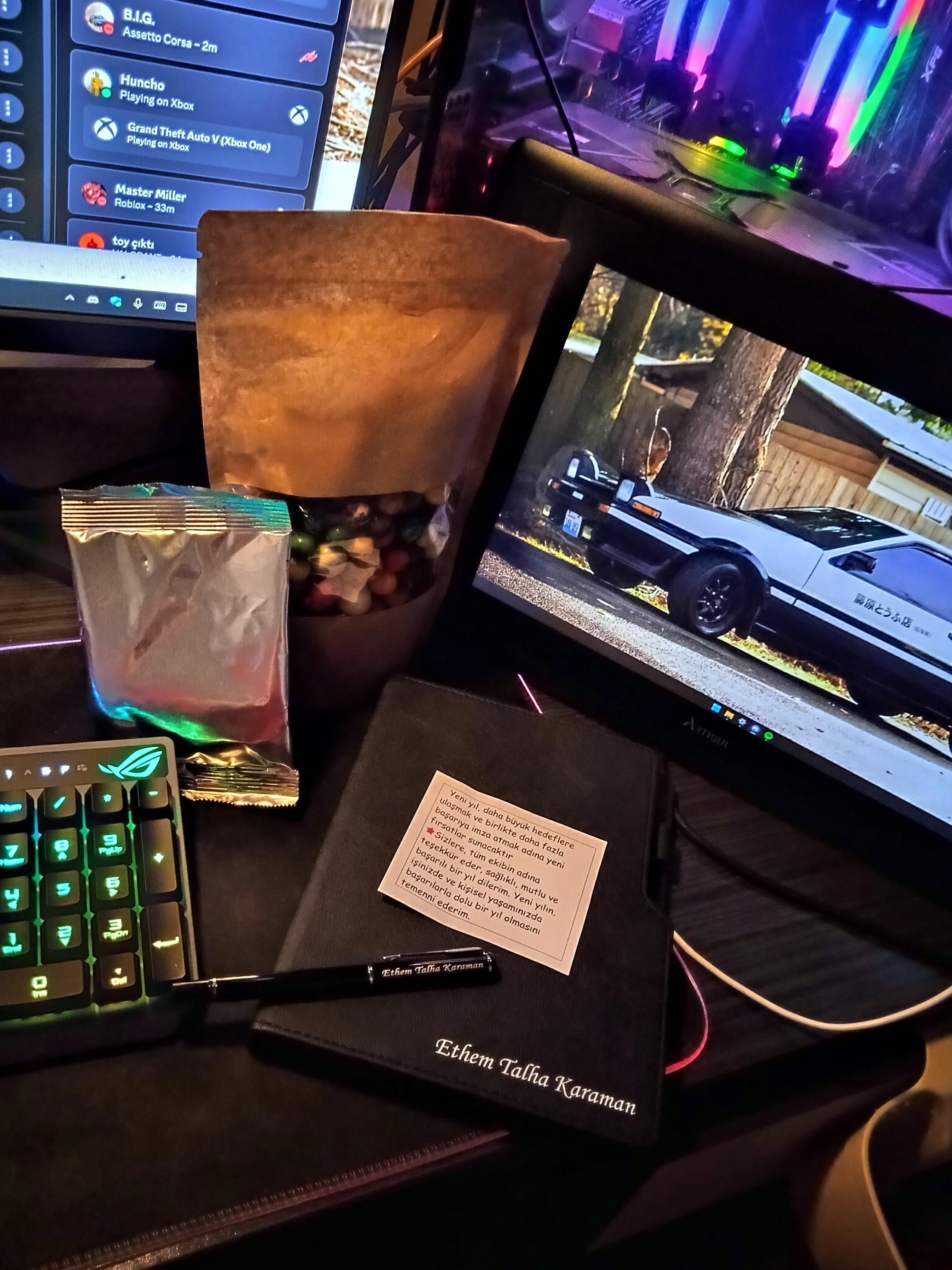 A computer desk setup with a monitor, a keyboard, a pen, a paper with writing, a brown bag of trail mix, and a smaller silver packet in front of a large computer monitor displaying a car. The monitor on the right shows an outdoor scene with a white and black car parked next to trees and a fence.