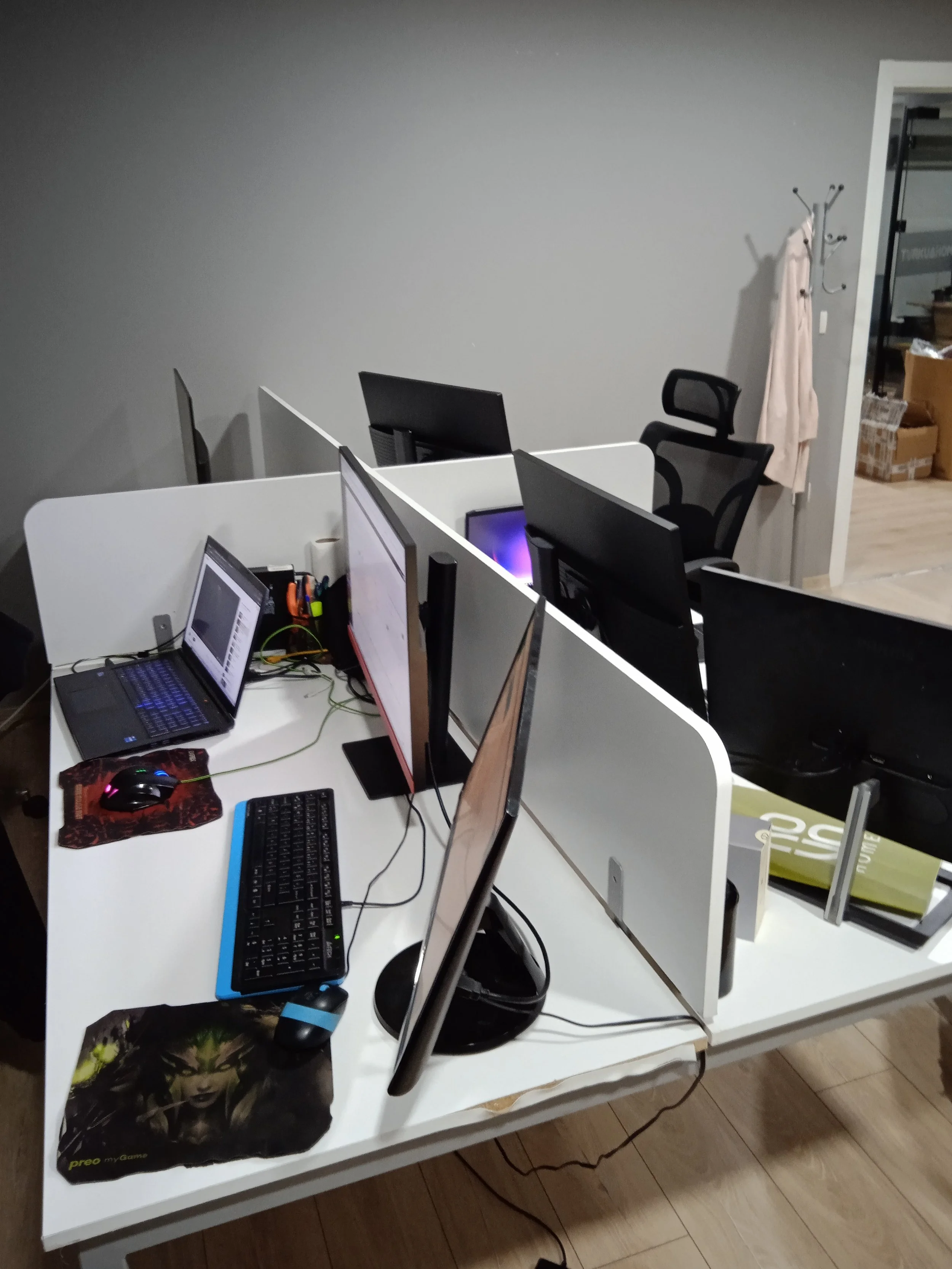 A white desk with multiple computer monitors, a laptop, and various office supplies. There are black office chairs and a coat rack with a garment hanging on it. The background is a plain gray wall.