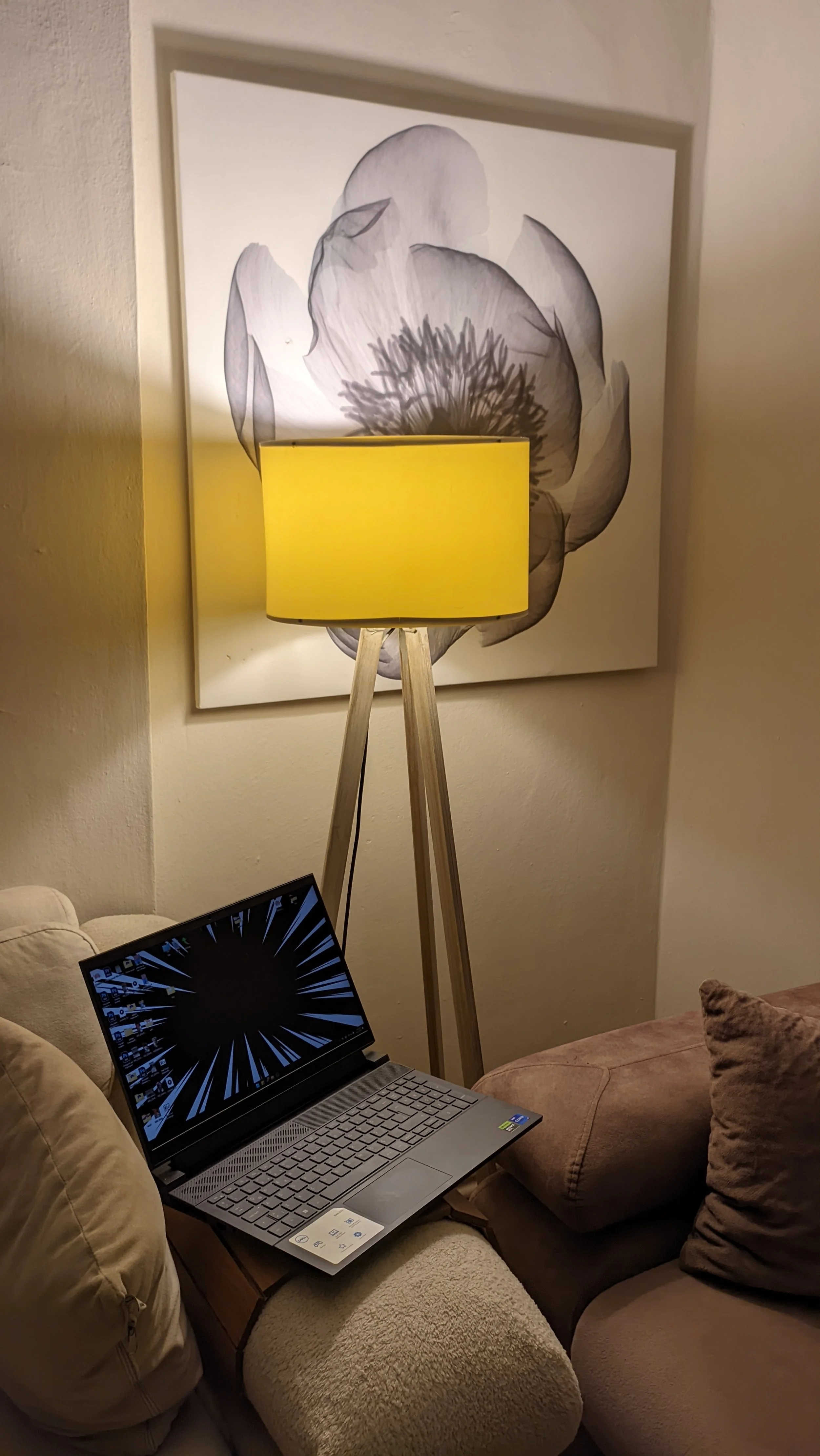 A room corner with a plaid armchair, a beige loveseat, a tall wooden tripod floor lamp with a yellow shade, a large black-and-white flower artwork on the wall, and a laptop on a small side table.