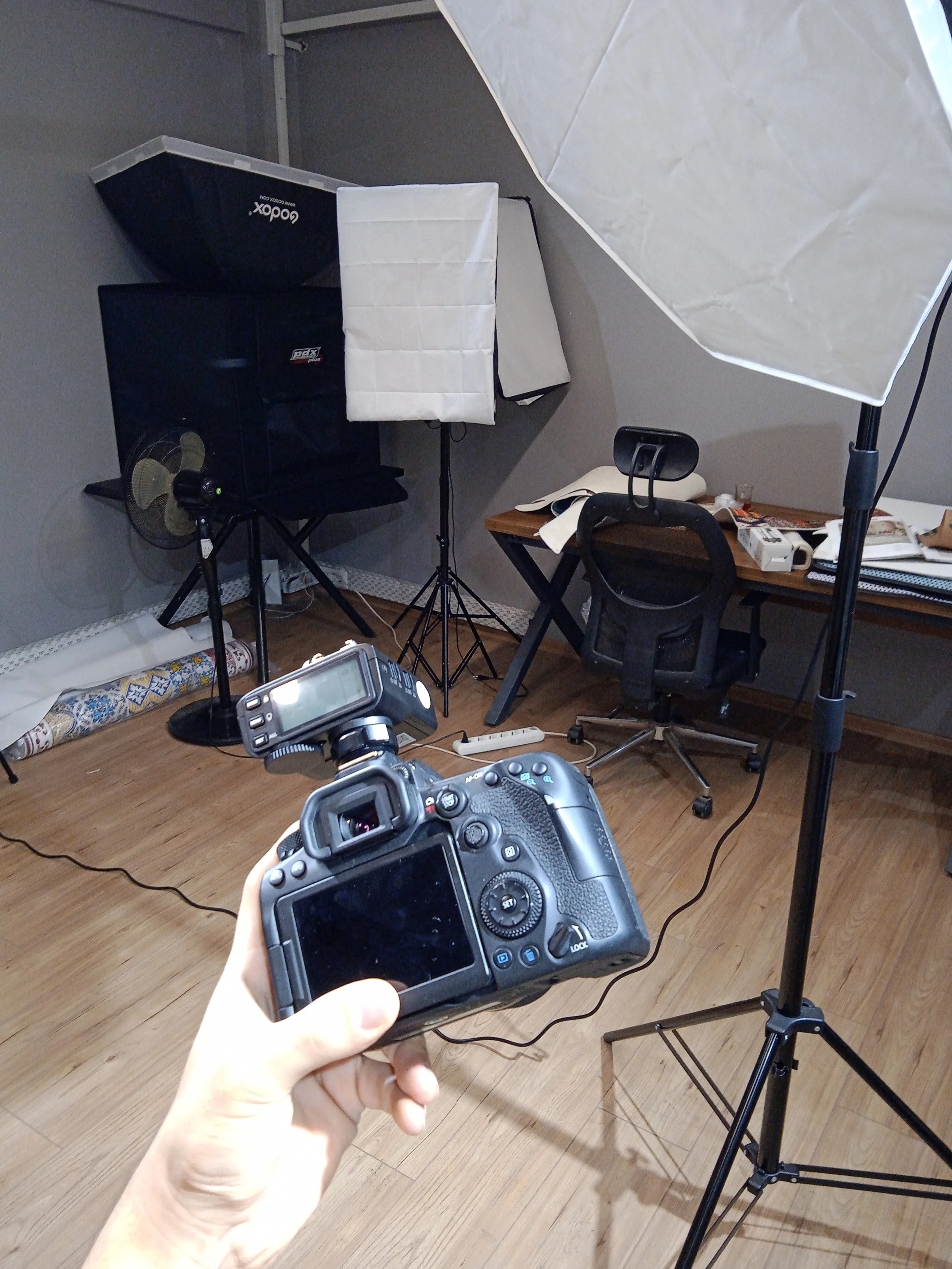 A person holding a DSLR camera in a photography studio setting with studio lights, a reflector, and a table with various items.