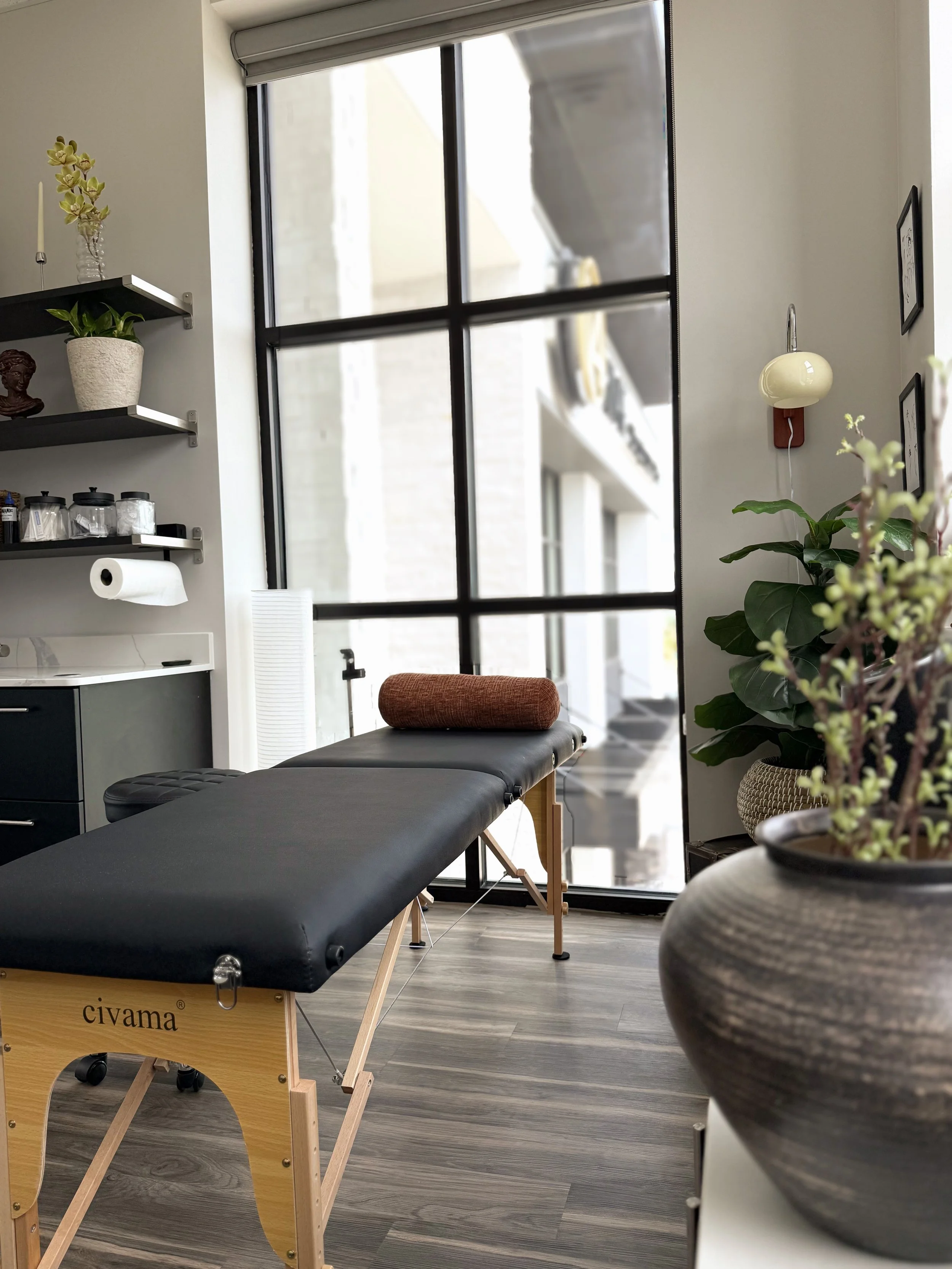 Interior of a room with a massage table, large windows, indoor plants, wall art, and a standing lamp.