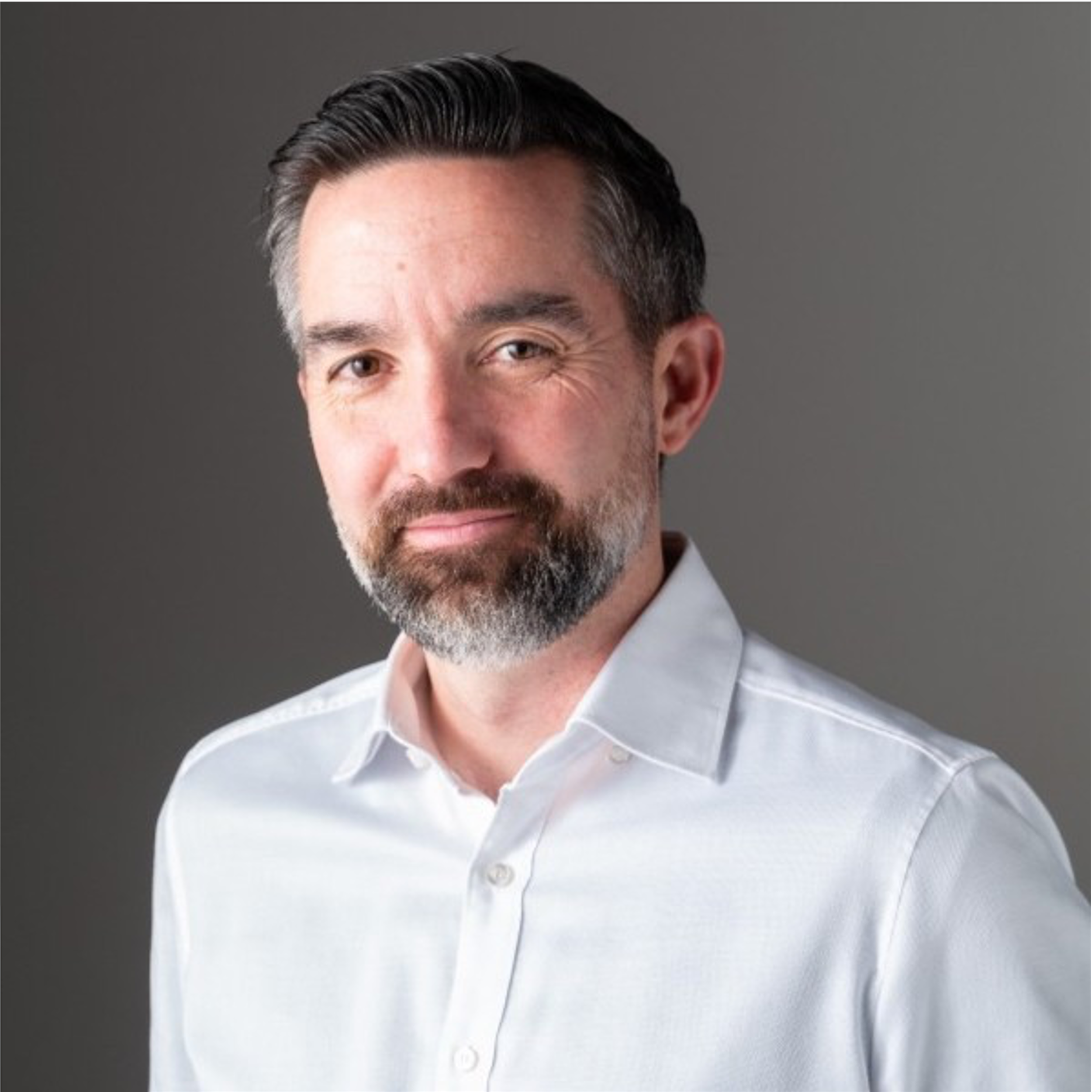 Close-up of a man wearing a light blue dress shirt with top buttons undone, featuring dark hair styled back and gray beard, against a gray background.
