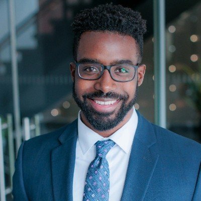 A man with dark curly hair, glasses, and a beard smiling in business attire, standing outside a modern building.