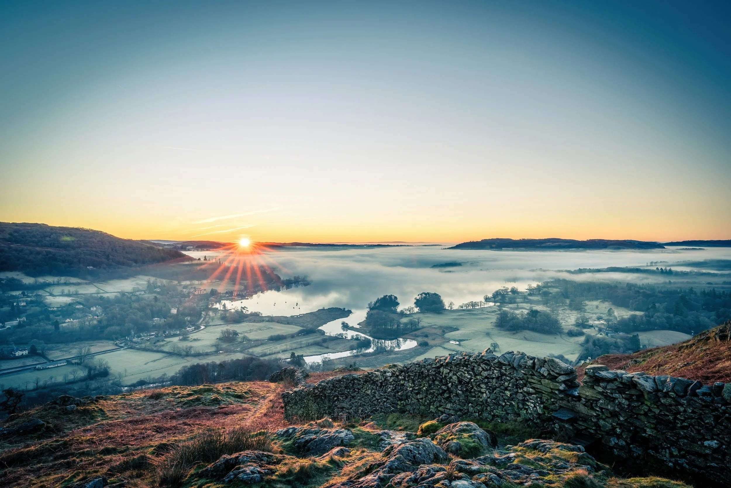 Sunrise over a foggy river valley with rolling hills and scattered trees — the Inversion Leadership metaphor for clarity above complexity