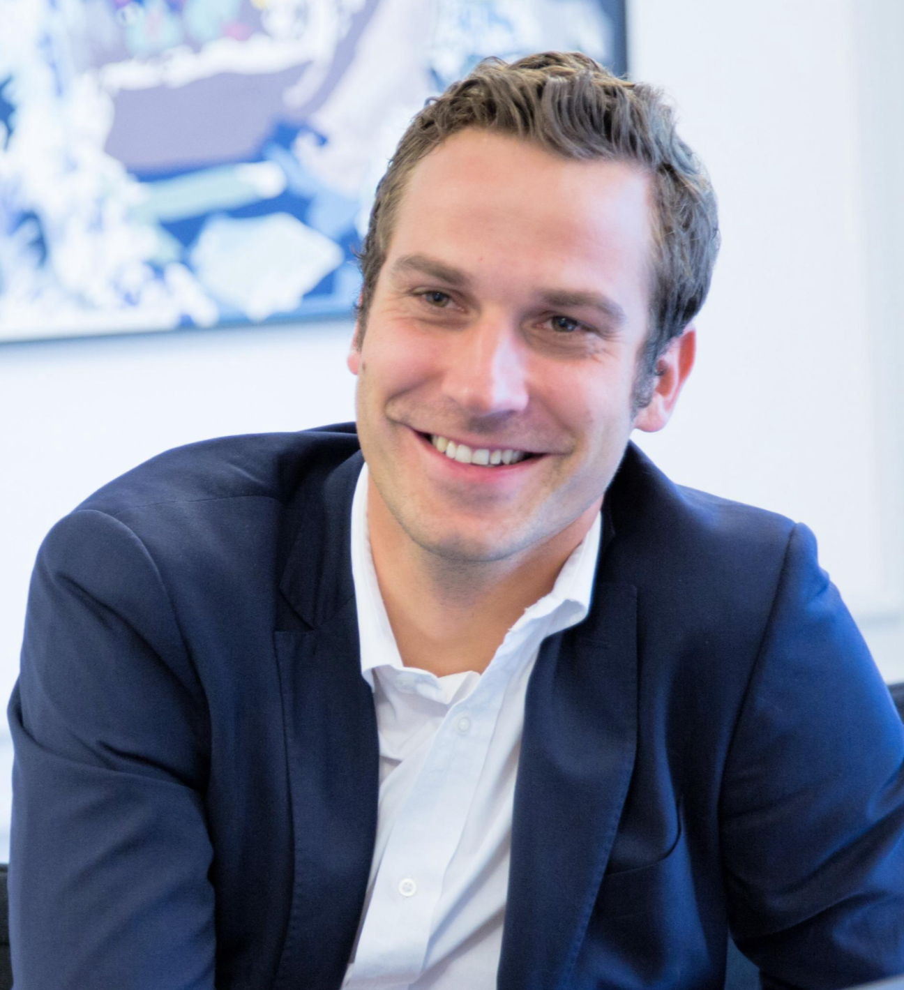 A smiling man in a navy blue suit and white shirt, sitting in an office with a blurred abstract painting in the background.