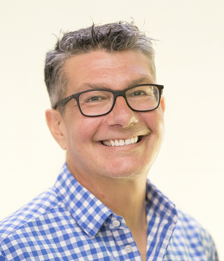 Smiling man with glasses and short gray hair, wearing a blue and white checkered shirt, against a light background.