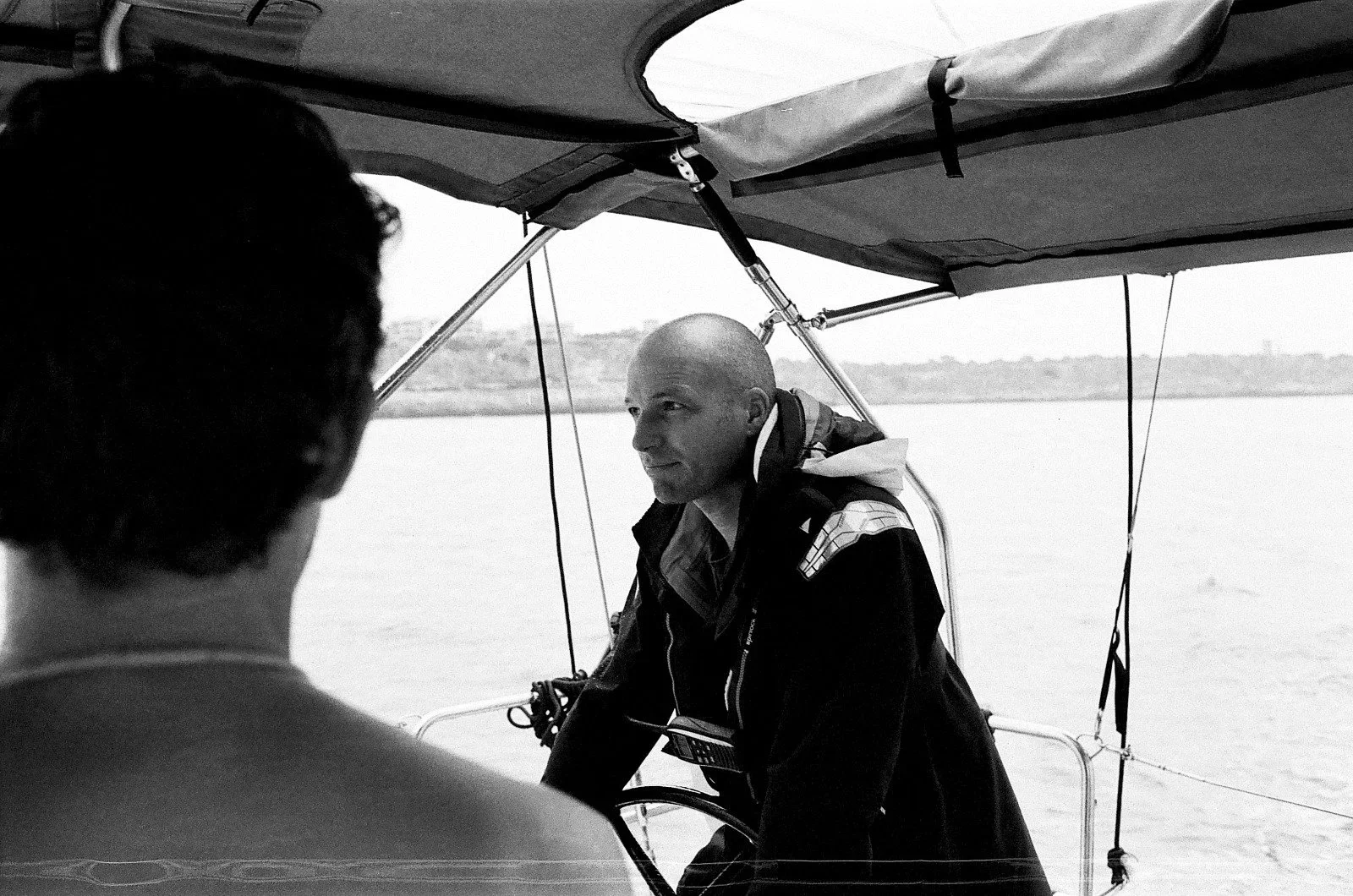 A man with a shaved head sitting on a boat, looking to the right, as another person faces away from the camera, with water and land in the background.