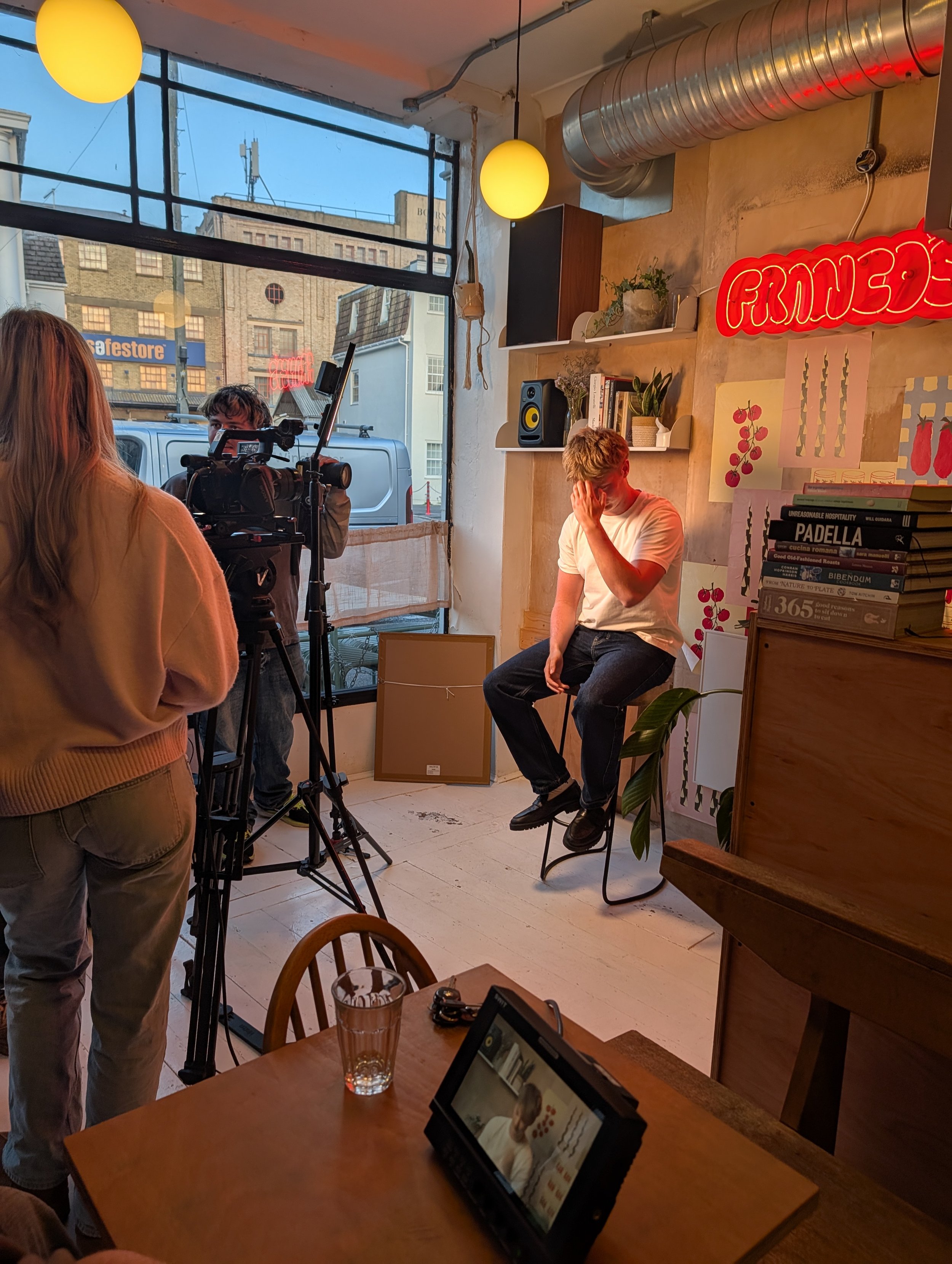 A man sitting on a high stool in a cafe setting, with a camera crew filming him. The cafe has large windows, yellow pendant lights, and a neon red sign Books and plants decorate the shelves on the wall. A tablet or digital device displaying an image and text is on a wooden table in the foreground, along with a pint glass and some accessories.